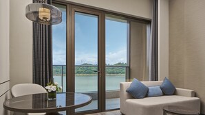 A modern hotel room with a large sliding glass door leading to a scenic view of a lake and hills. There is a round table with a vase of flowers, a sofa with blue cushions, and a small side table. - Crowne Plaza Ganzhou by IHG (Ganzhou)
