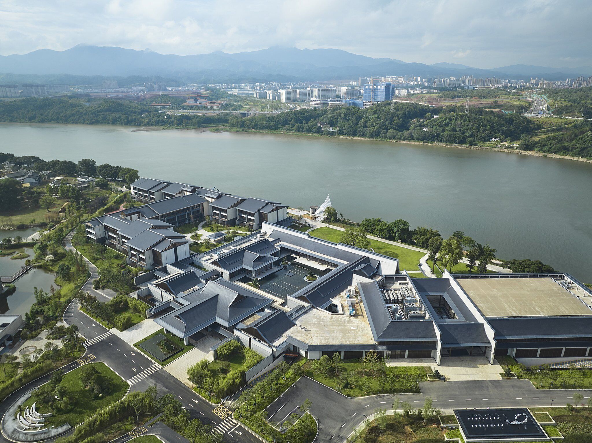 Vue aérienne d’un complexe immobilier moderne composé de plusieurs bâtiments, avec des aires de stationnement et une rivière en arrière-plan.
