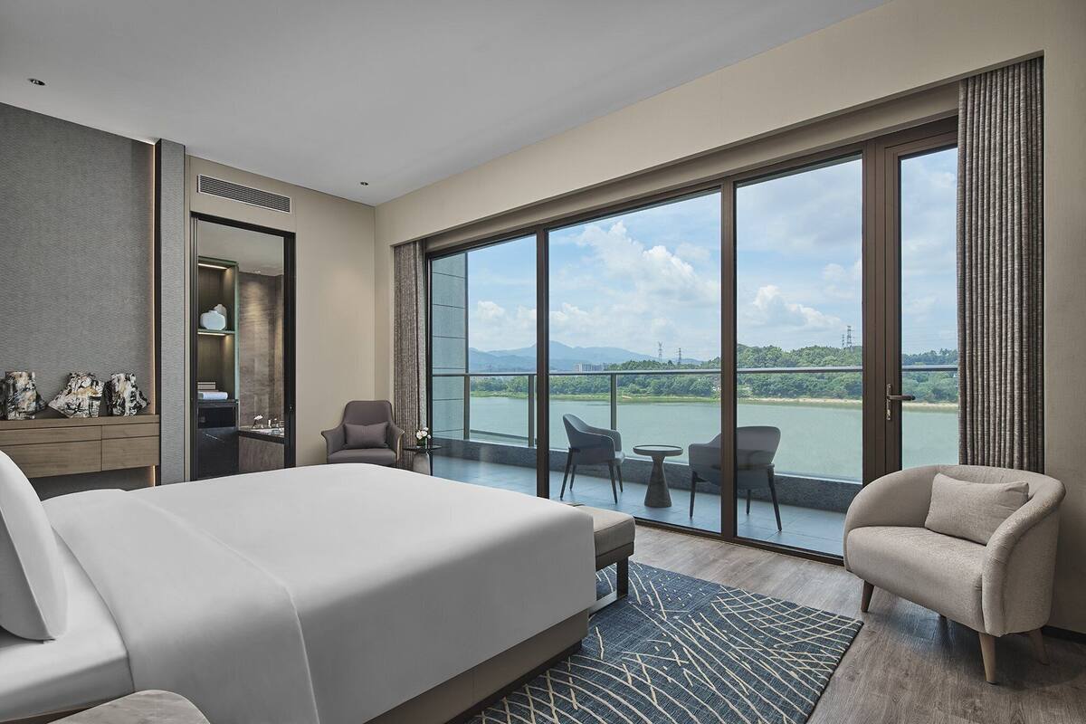 Ein Hotelzimmer mit einem großen Bett, einer Sitzecke mit Stühlen, einem kleinen Tisch und Blick auf ein Gewässer durch große Fenster.