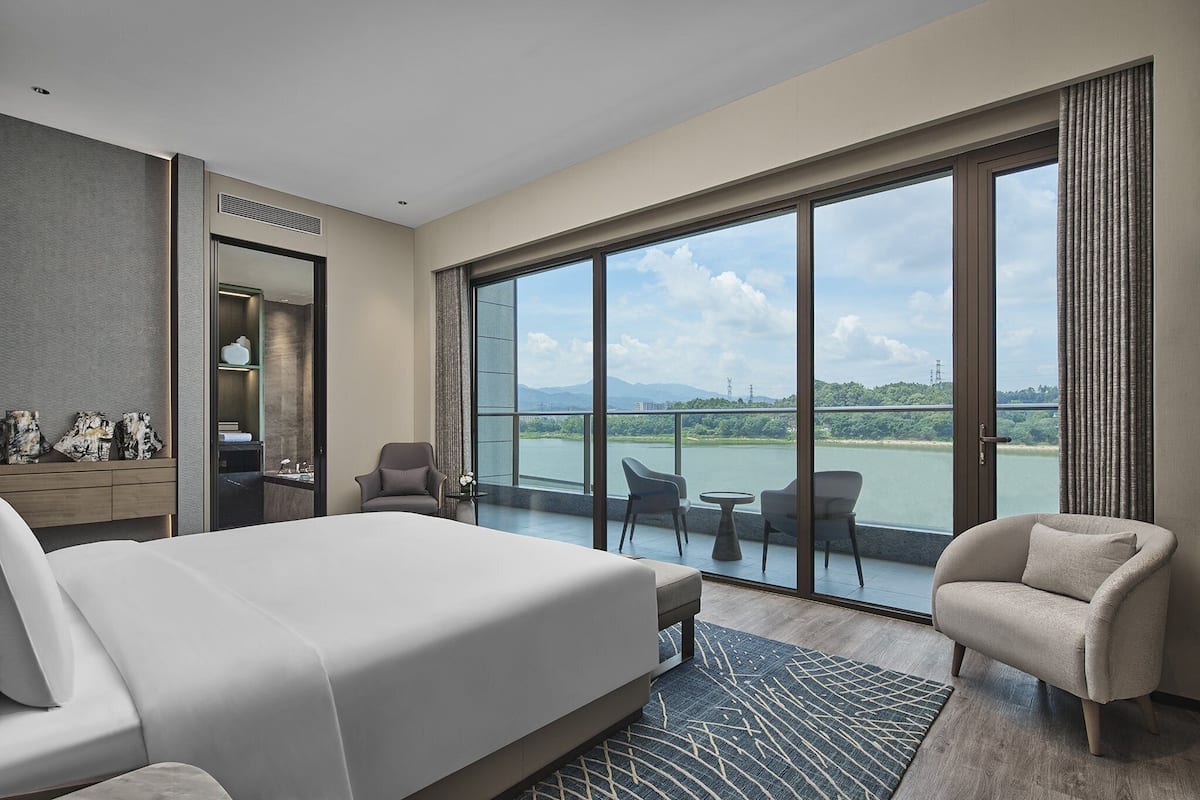 A hotel room with a large bed, a seating area with chairs, a small table, and a view of a body of water through large windows.