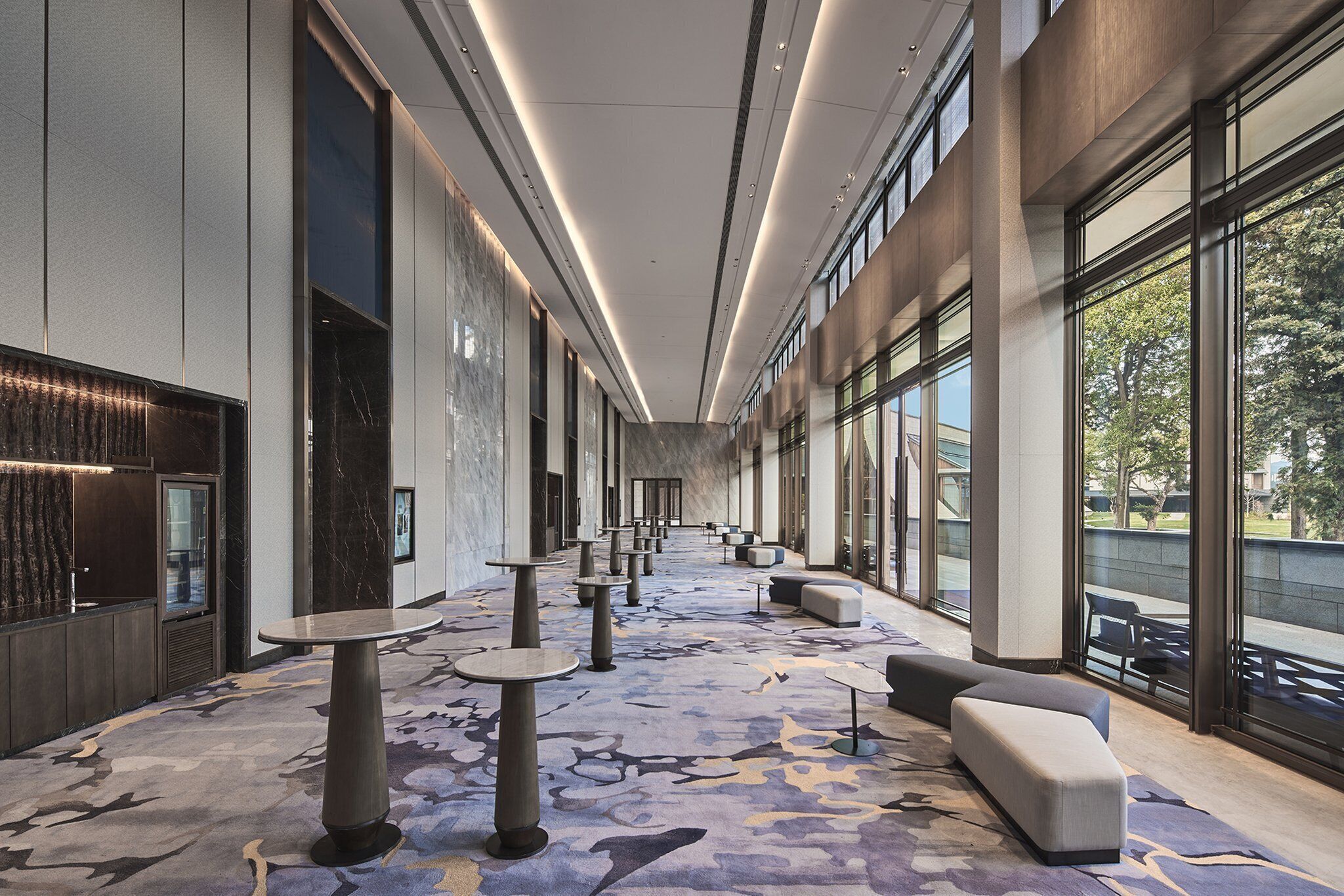 A spacious corridor with round tables and seating areas, large windows offering views of the outdoors, and a modern interior design.