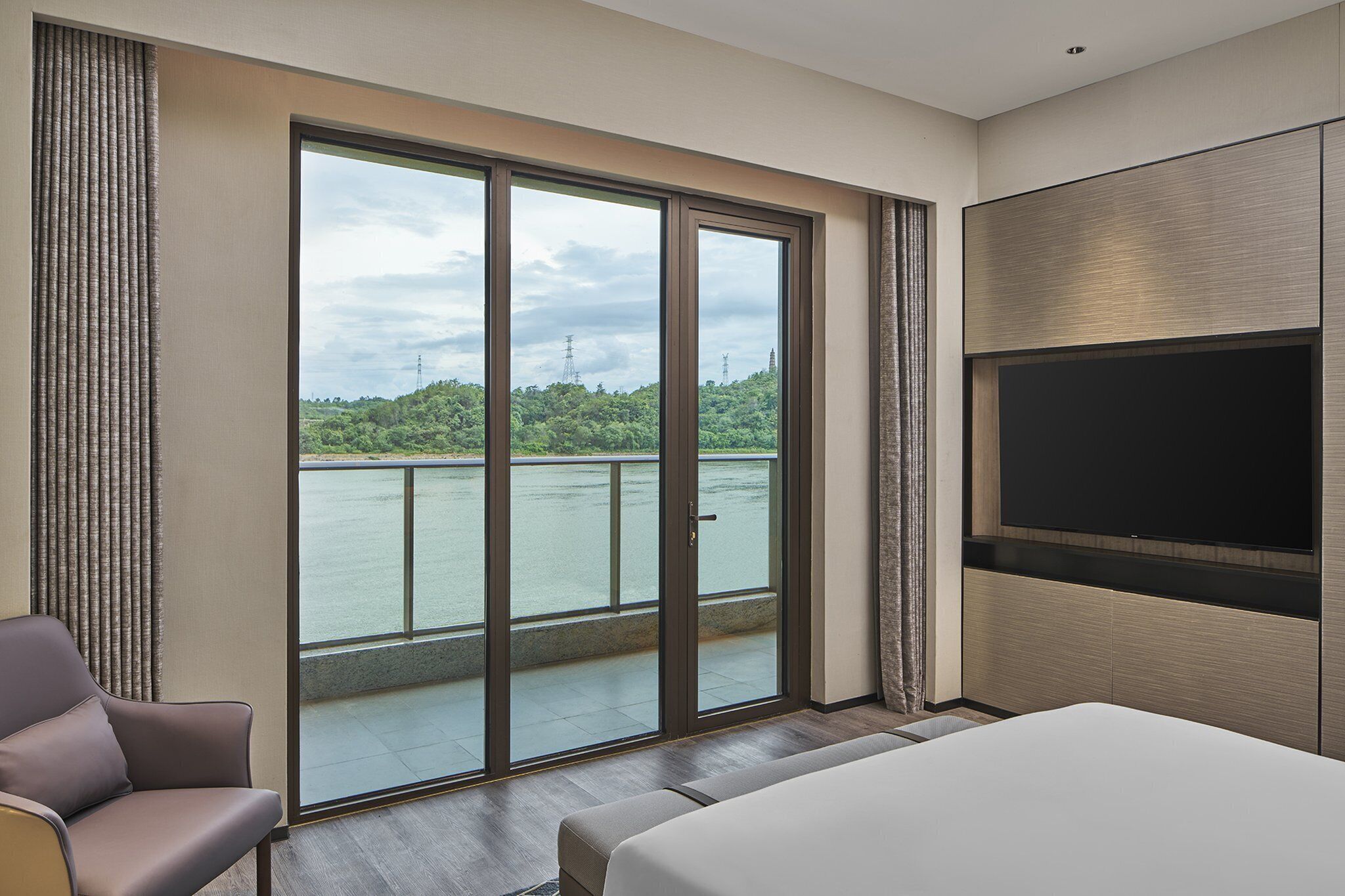 A hotel room with a large sliding glass door leading to a balcony with a view of a body of water and greenery. There is a bed, a chair, and a flat-screen TV mounted on the wall.