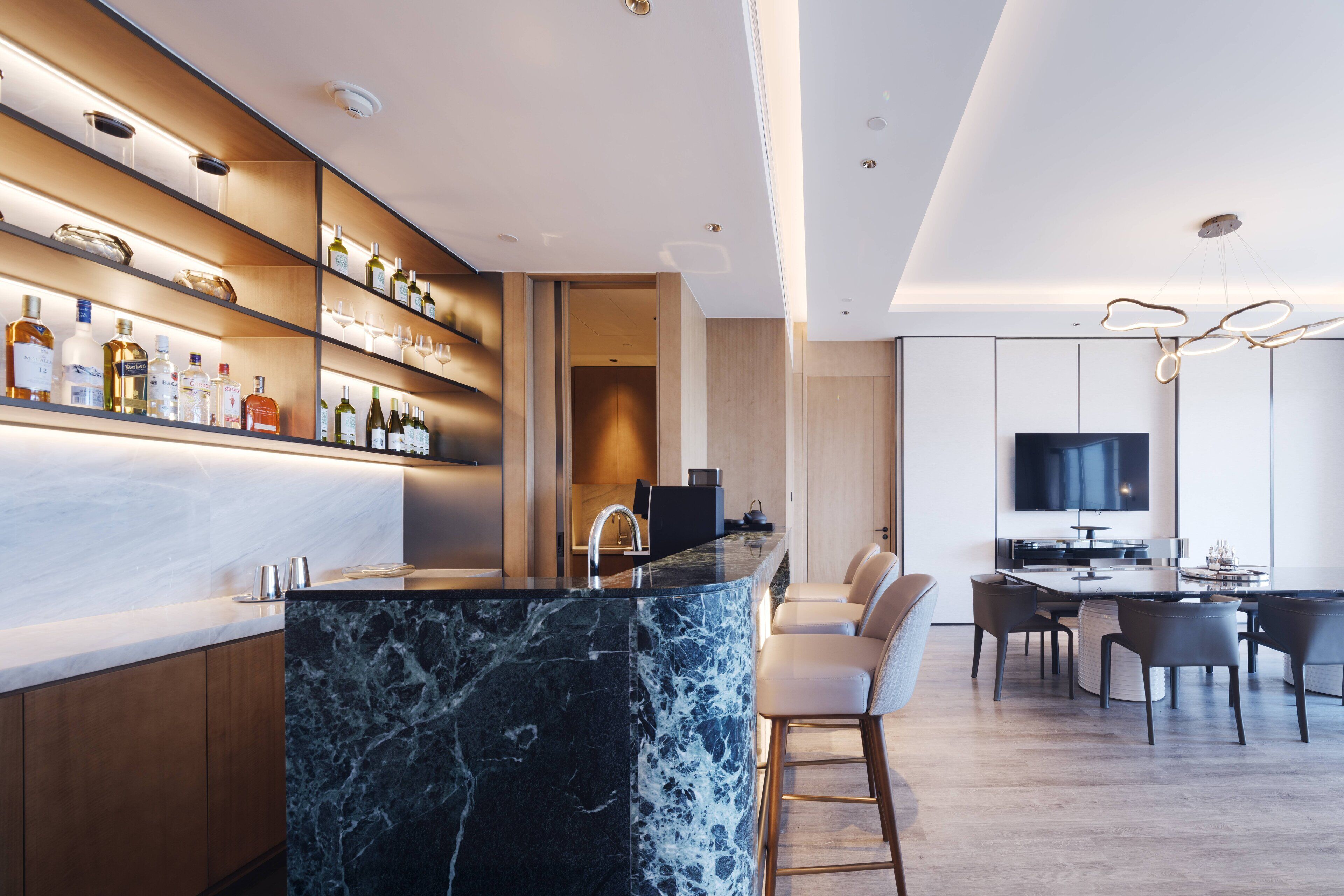 A modern kitchen with a marble island, built-in shelves with bottles, and a dining area with a TV.