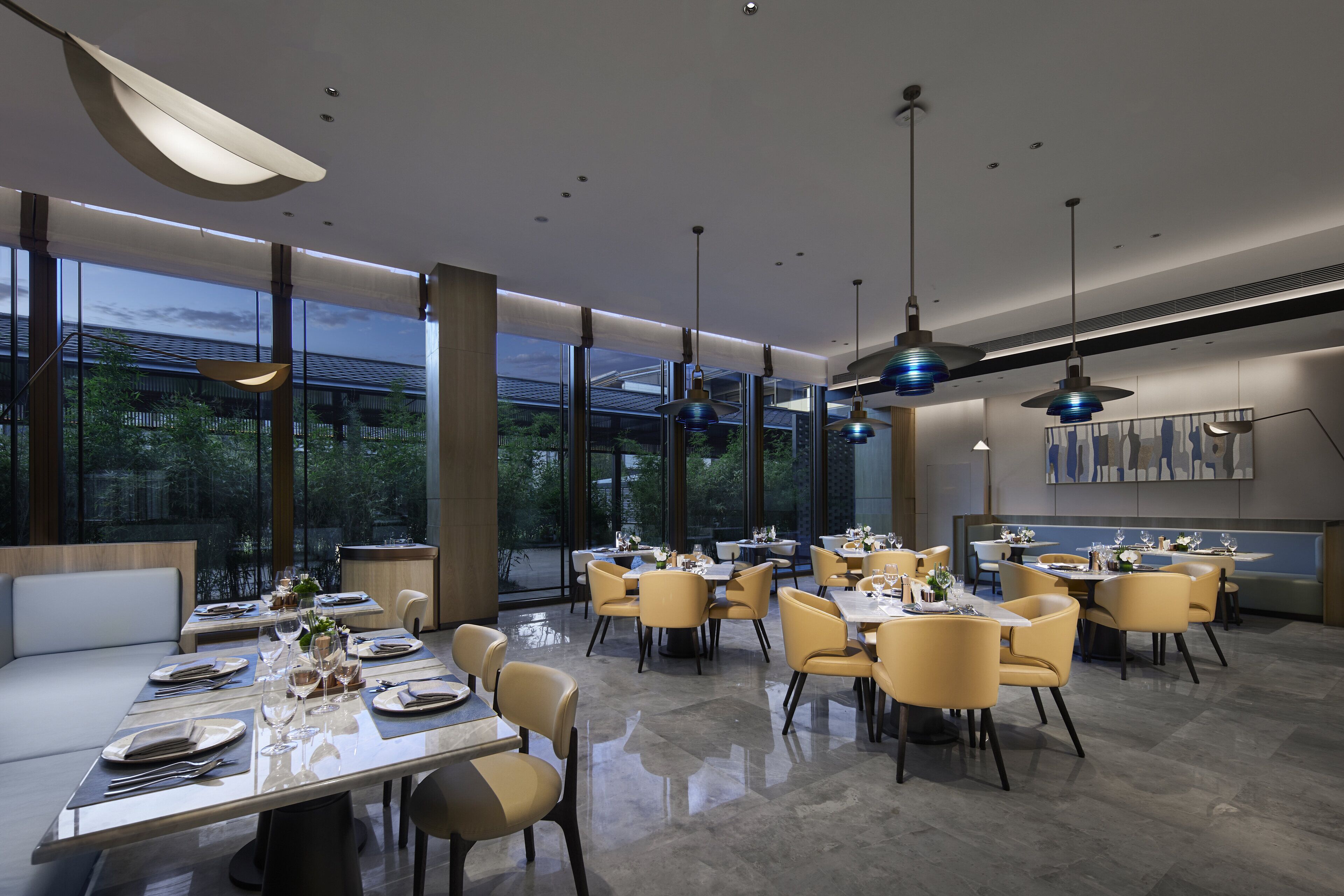 Un comedor moderno con varias mesas y sillas, ventanas amplias e iluminación colgante.
