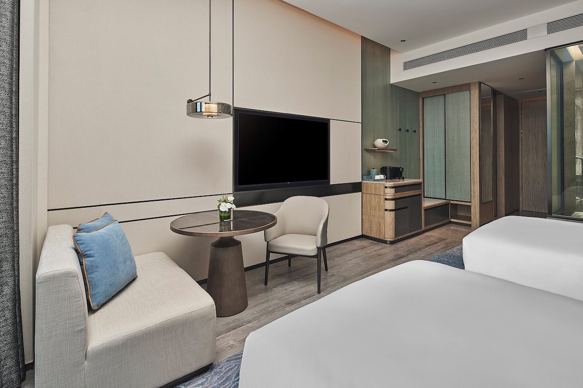 Habitación de hotel moderna con cama, sofá, mesita redonda pequeña, silla, televisor, minibar y cabina de ducha de vidrio.