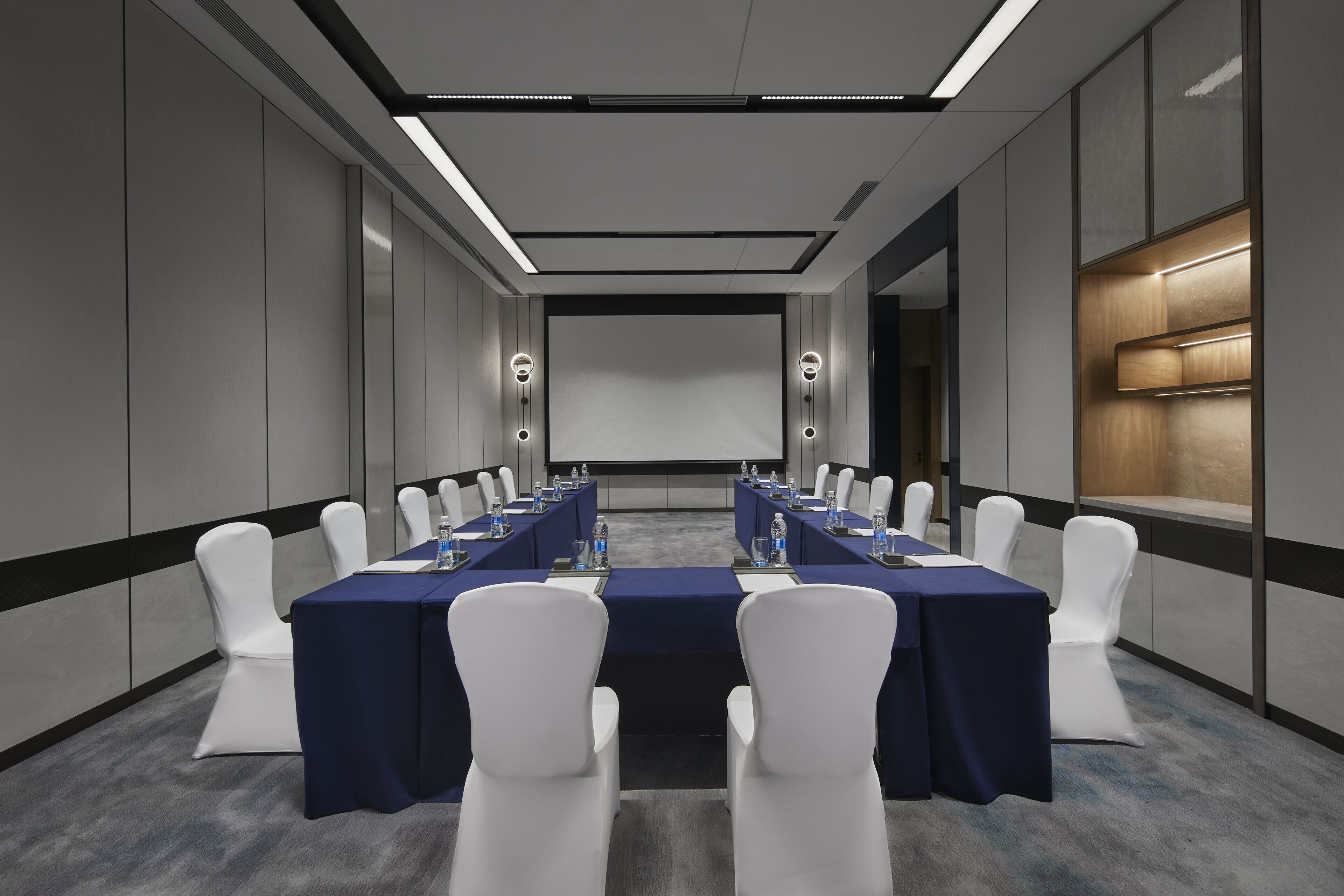 A conference room with a long table, white chairs, and a projector screen.