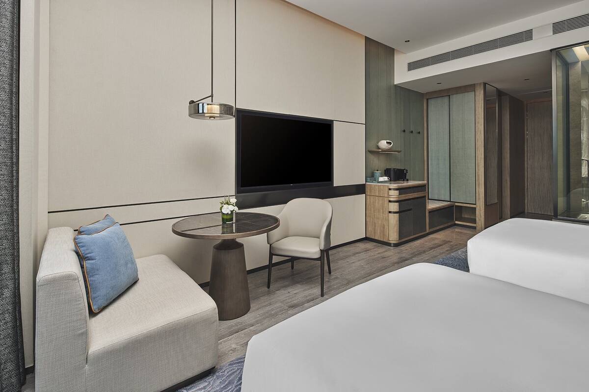 Ein modernes Hotelzimmer mit Sofa, rundem Tisch, Stuhl, Bett, Fernseher und Minibar.