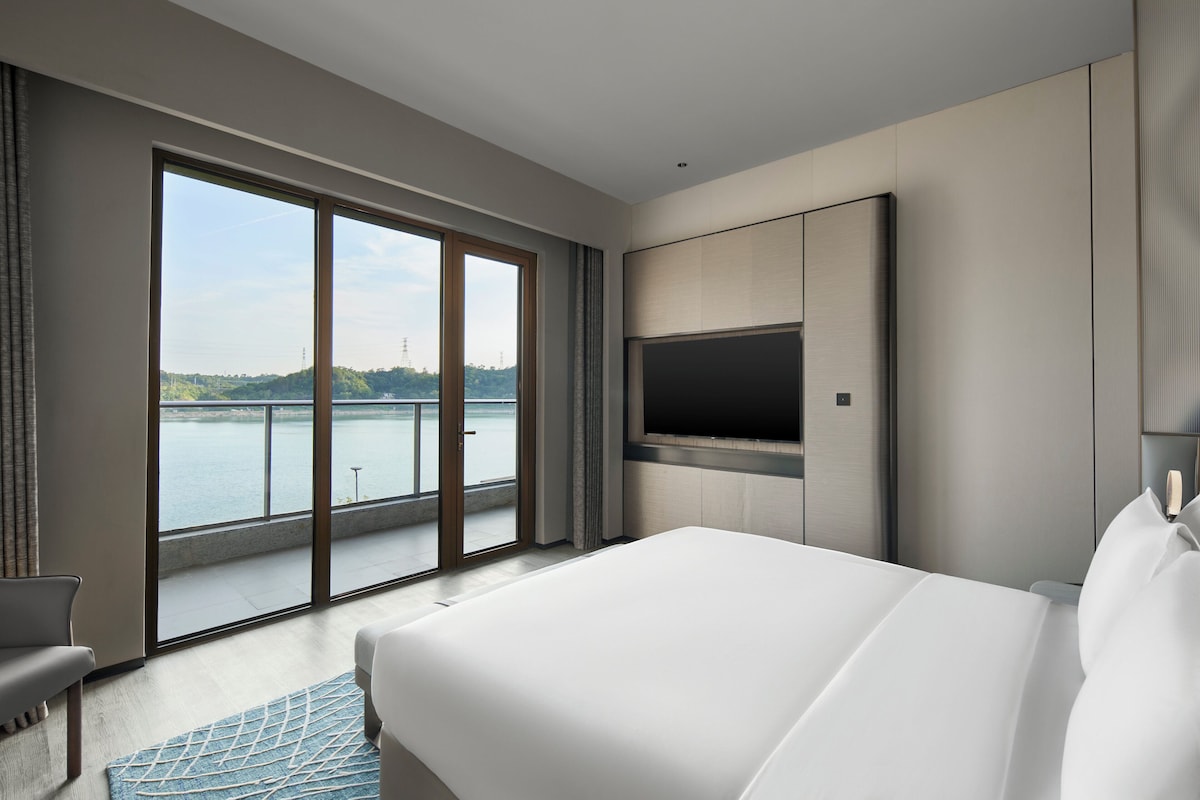 Habitación de hotel con una cama grande, televisor de pantalla plana montado en la pared y puerta corrediza de vidrio que da a un balcón con vistas a una masa de agua y vegetación.