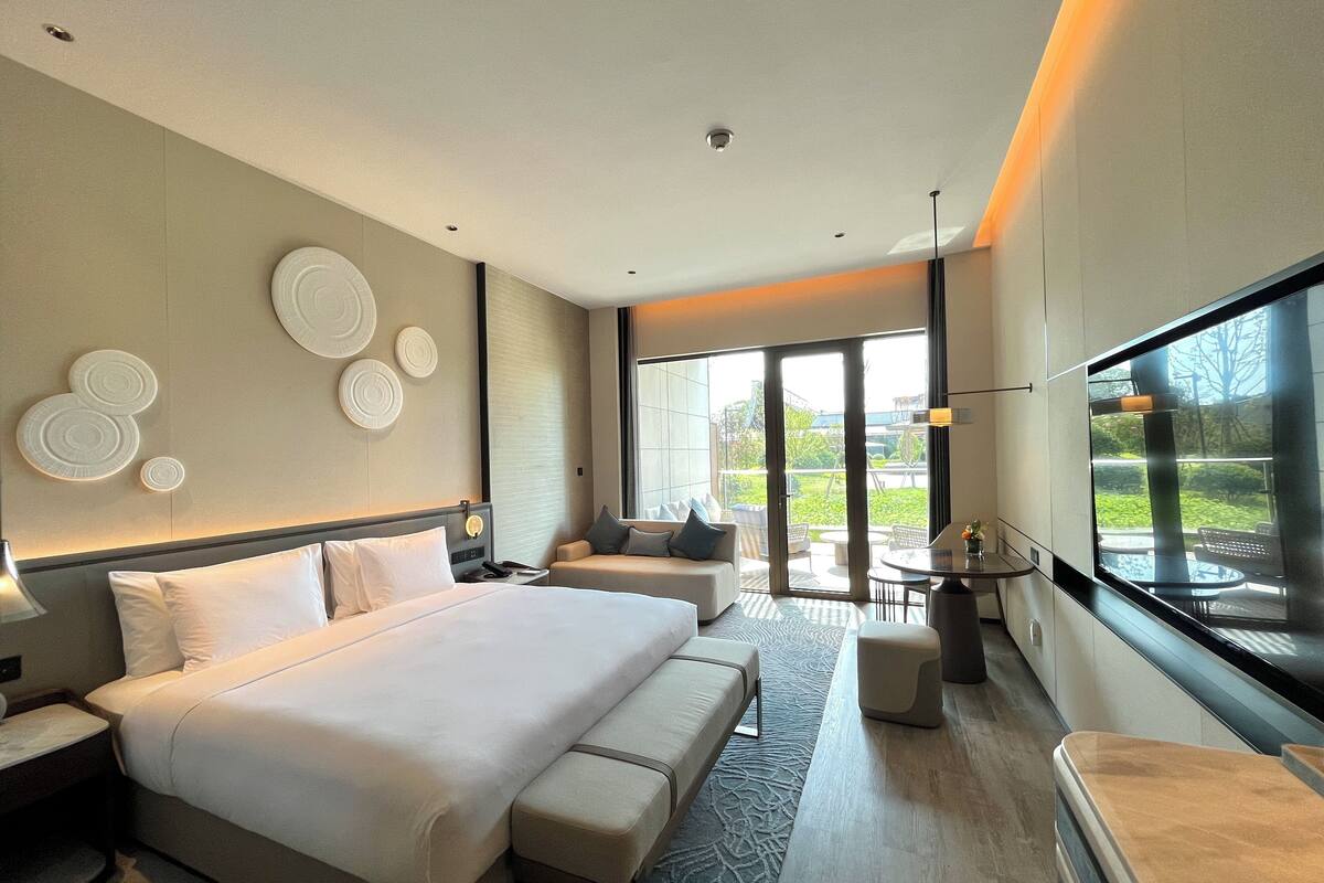 Ein modernes Hotelzimmer mit einem großen Bett, einer Couch, einem Esstisch und Blick ins Freie.