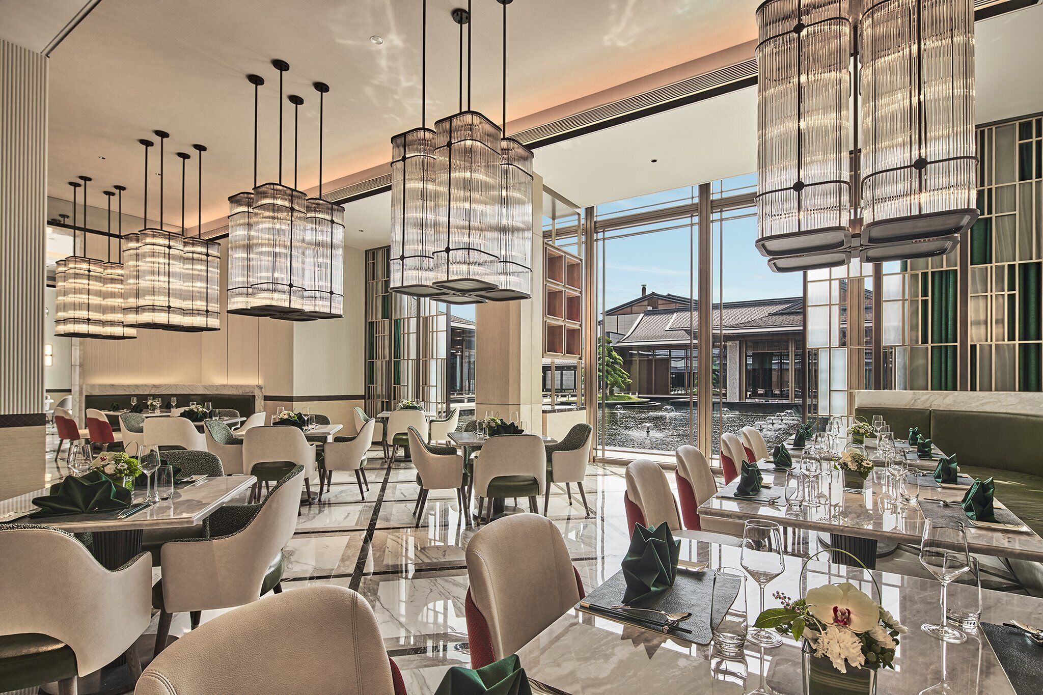 A well-lit dining area with multiple tables set for guests, featuring modern chandeliers and large windows offering a view of the outside.