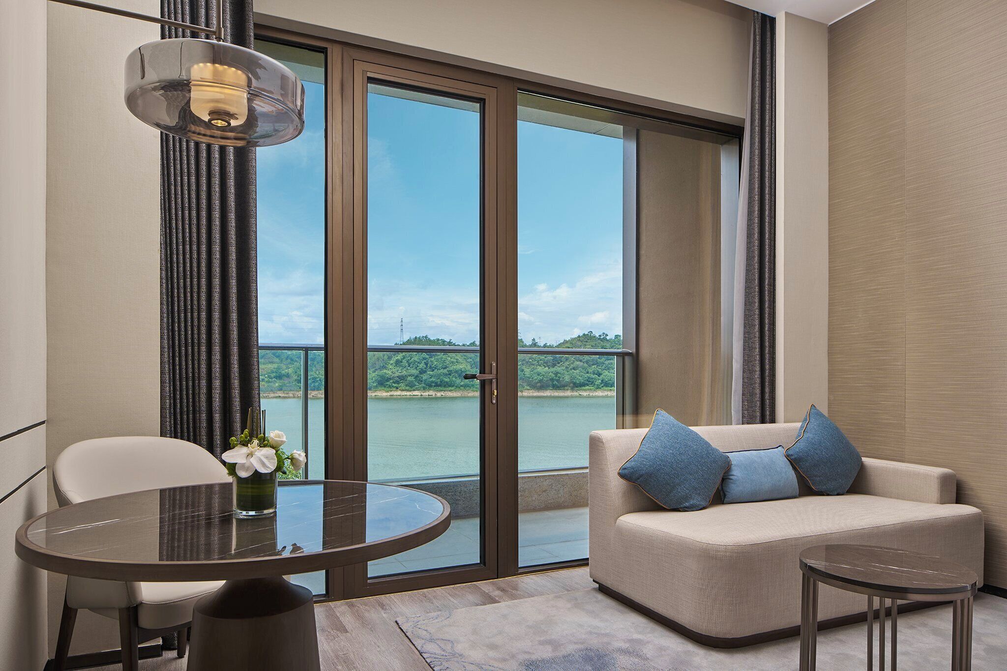 Una habitación moderna con una puerta de cristal que da a un balcón, una mesa redonda, un sofá con cojines y una lámpara colgante.