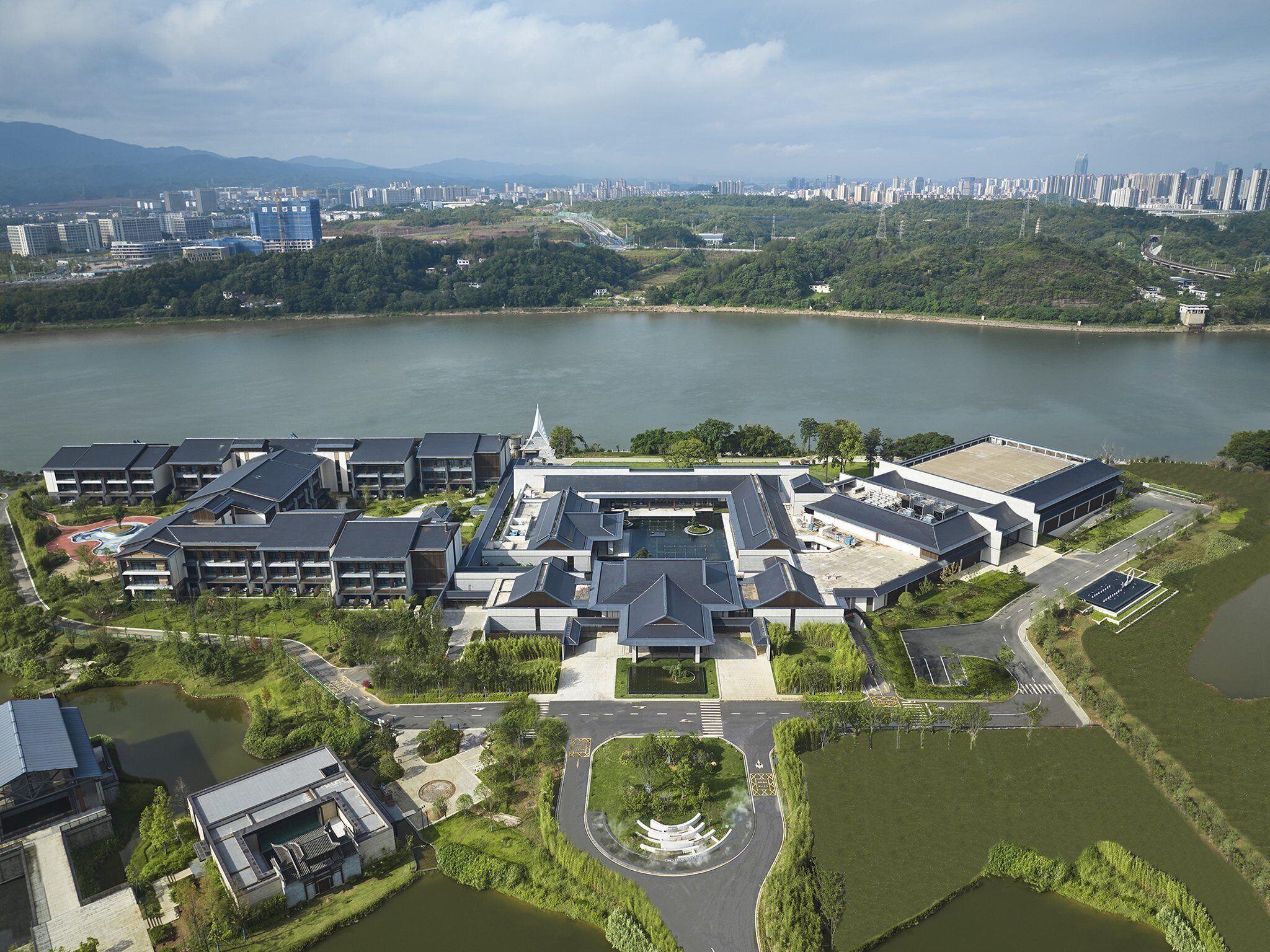 An aerial view of a modern hotel complex with multiple buildings, a central courtyard, and a lake in the background.