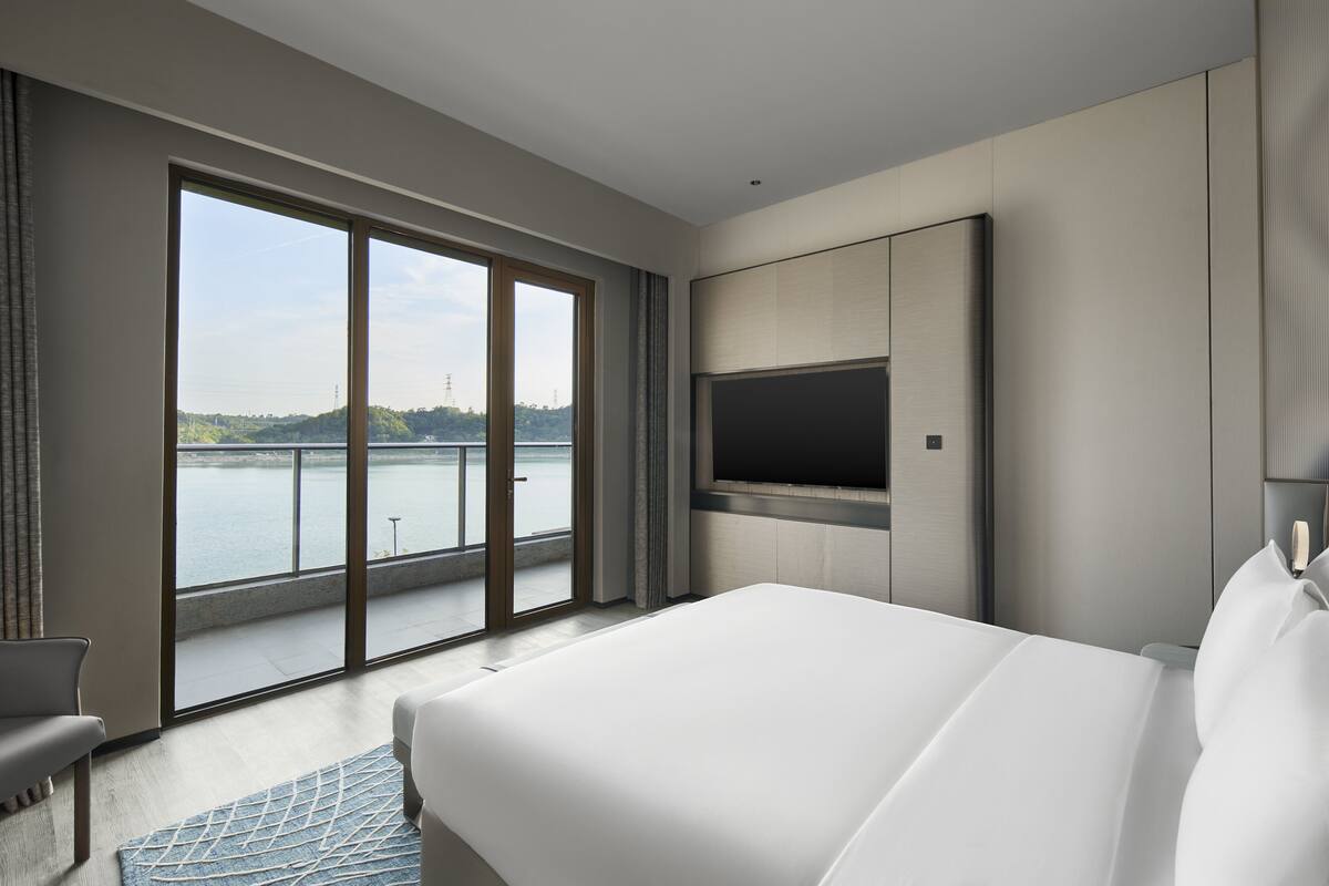 Ein modernes Hotelzimmer mit einem großen Bett, einem flachbildfernseher an der Wand und einer Schiebetür zu einem Balkon mit Blick auf ein Gewässer und viel Grün.