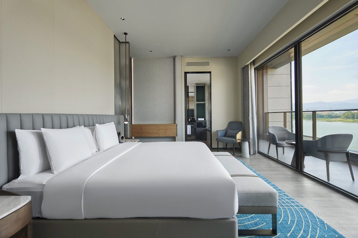 Una habitación de hotel moderna con una cama grande, una zona de estar con sillones y vistas al exterior, además de un balcón accesible mediante un cerramiento corredero.