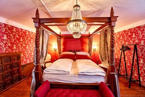 Ein Bett mit Baldachin, eine Chandelierleuchter, rote blumige Tapete und ein Holzboden.