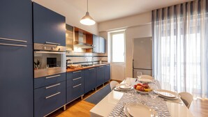 A modern kitchen with blue cabinets, a white dining table set with plates and glasses, and a window with sheer curtains. - BnButler - Strigelli 13 (Milan)