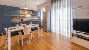 A modern kitchen with blue cabinets, a dining table with chairs, a refrigerator, and a flat-screen TV on a white stand. - BnButler - Strigelli 13 (Milan)