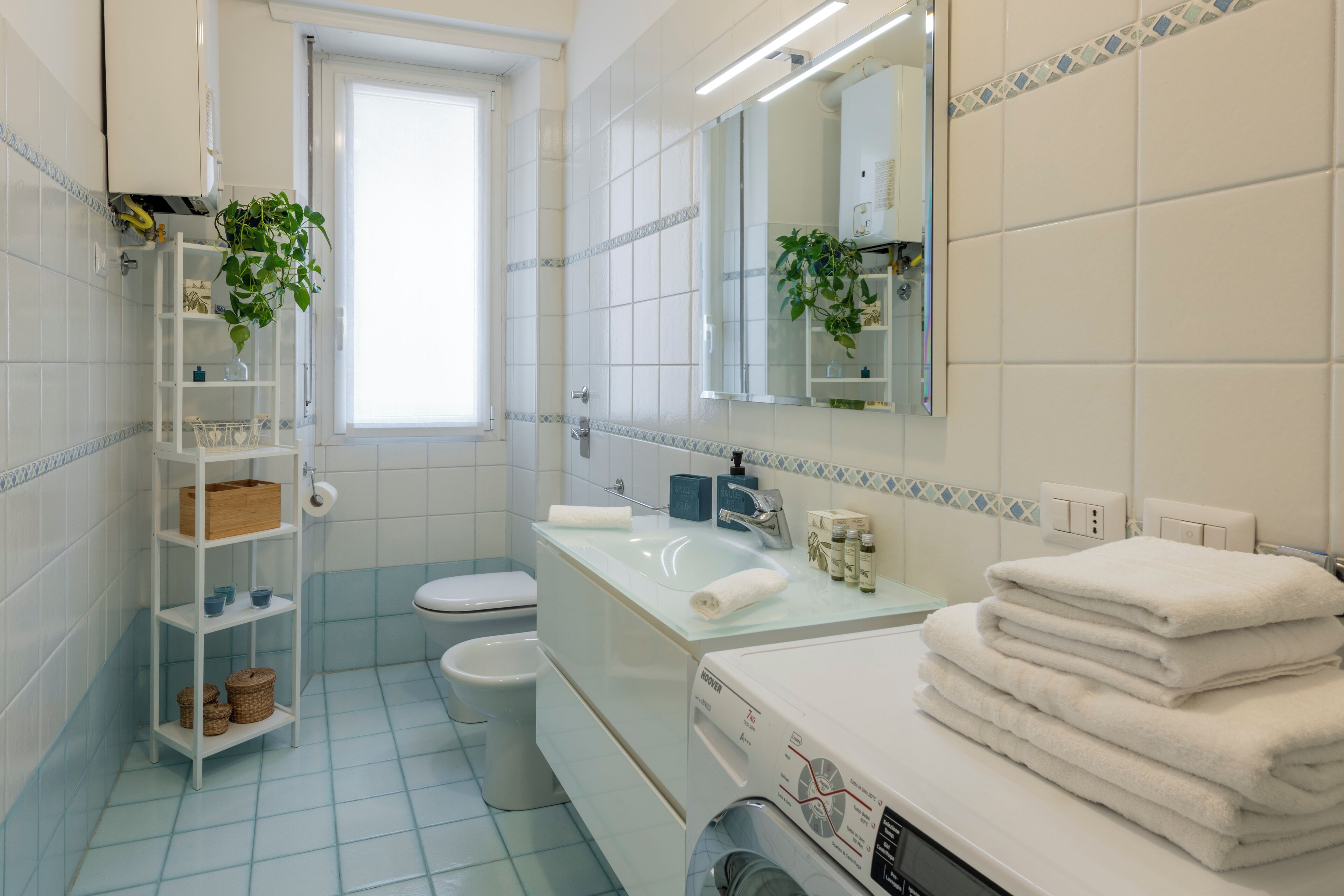 Un baño con lavamanos blanco, espejo, inodoro y estante con artículos de aseo.
