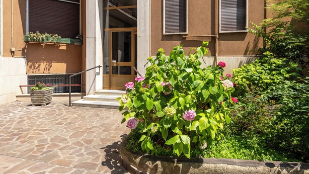 A stone-paved entrance to a building with a large wooden door, a window with a metal shutter, and a planter with flowers. - BnButler - Strigelli 13 (Milan)