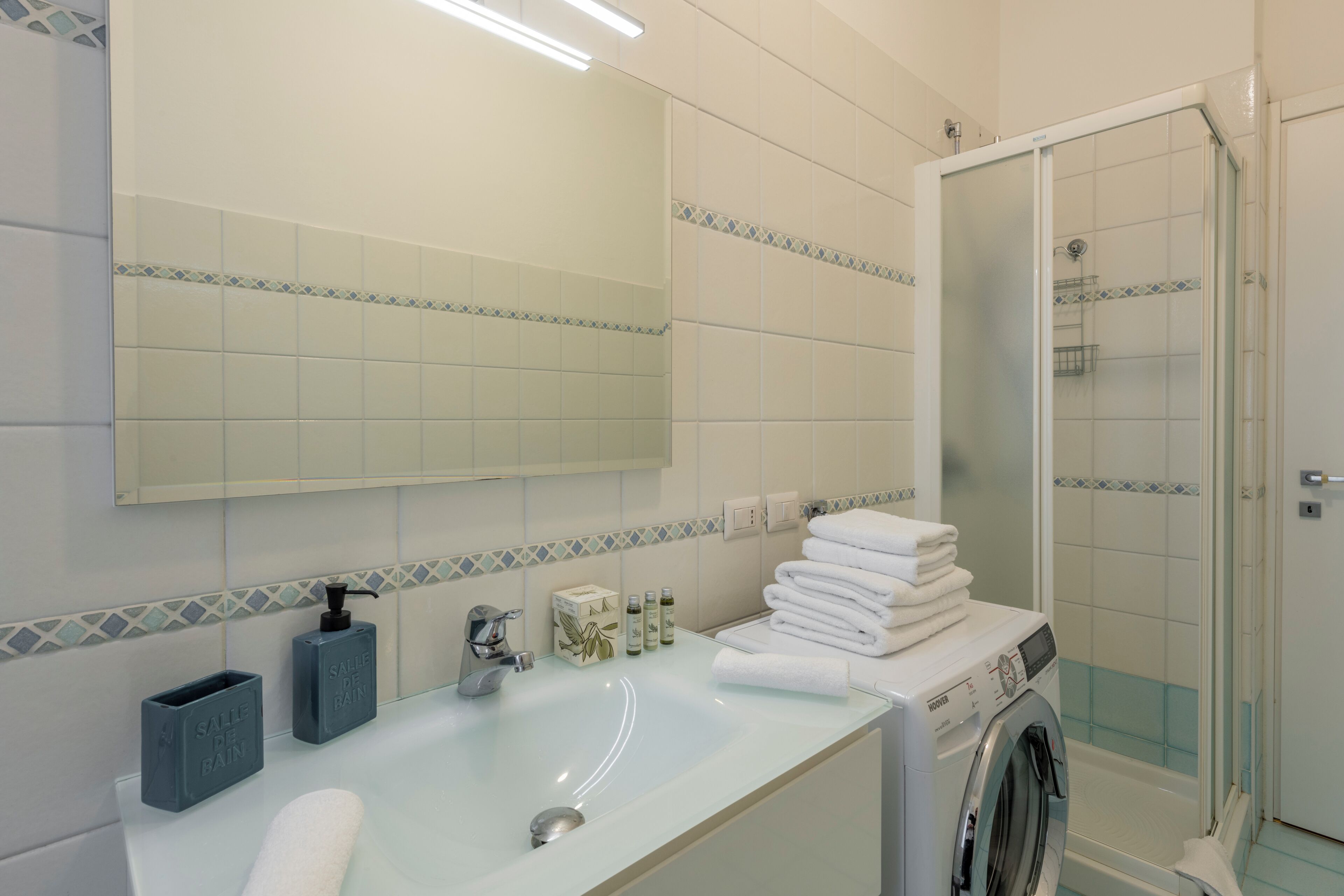 A bathroom with a sink, mirror, toiletries, stacked towels, and a washing machine.