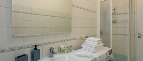 A bathroom with a sink, mirror, toiletries, stacked towels, and a washing machine.