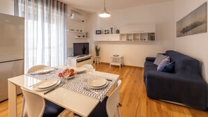 A modern living room with a dark blue sofa, a dining table set with plates and glasses, a TV, and a painting on the wall. - BnButler - Strigelli 13 (Milan)