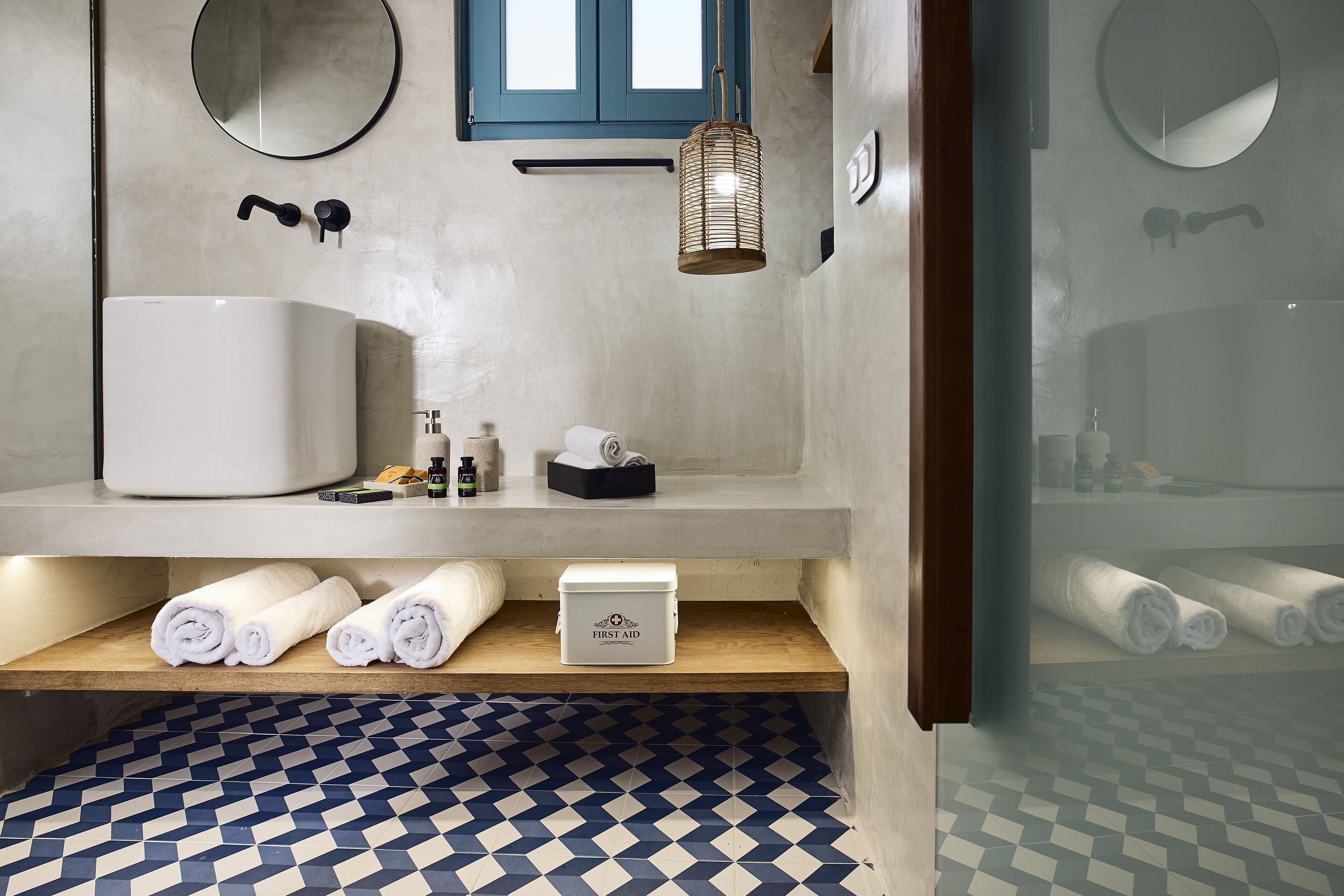 Un baño con un lavamanos grande de color blanco, un estante de madera con toallas, una caja de primeros auxilios y un suelo estampado.
