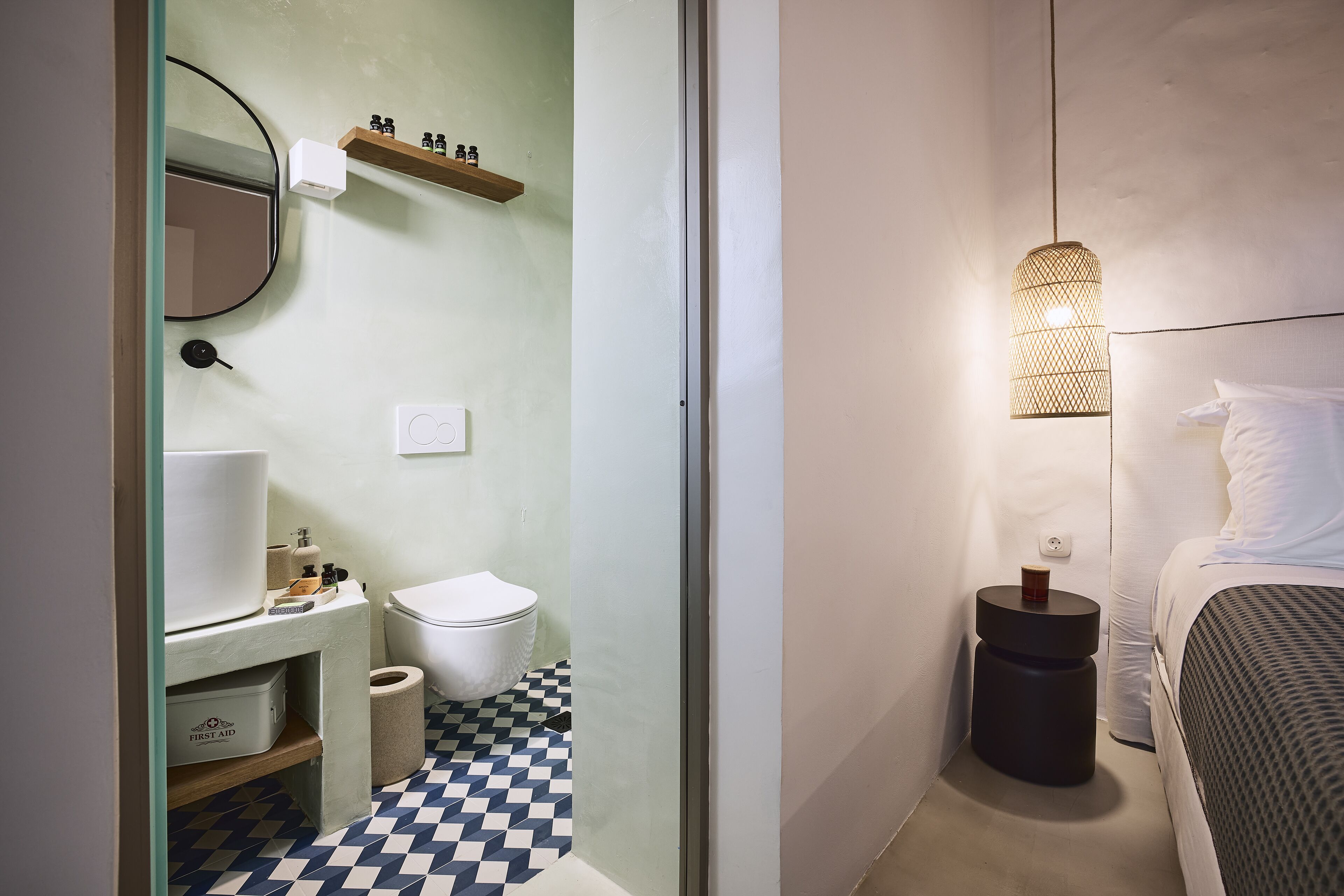 Une petite salle de bain comprenant des toilettes, un lavabo et un miroir. On y trouve une étagère avec des articles de toilette et un luminaire suspendu au-dessus du lit.