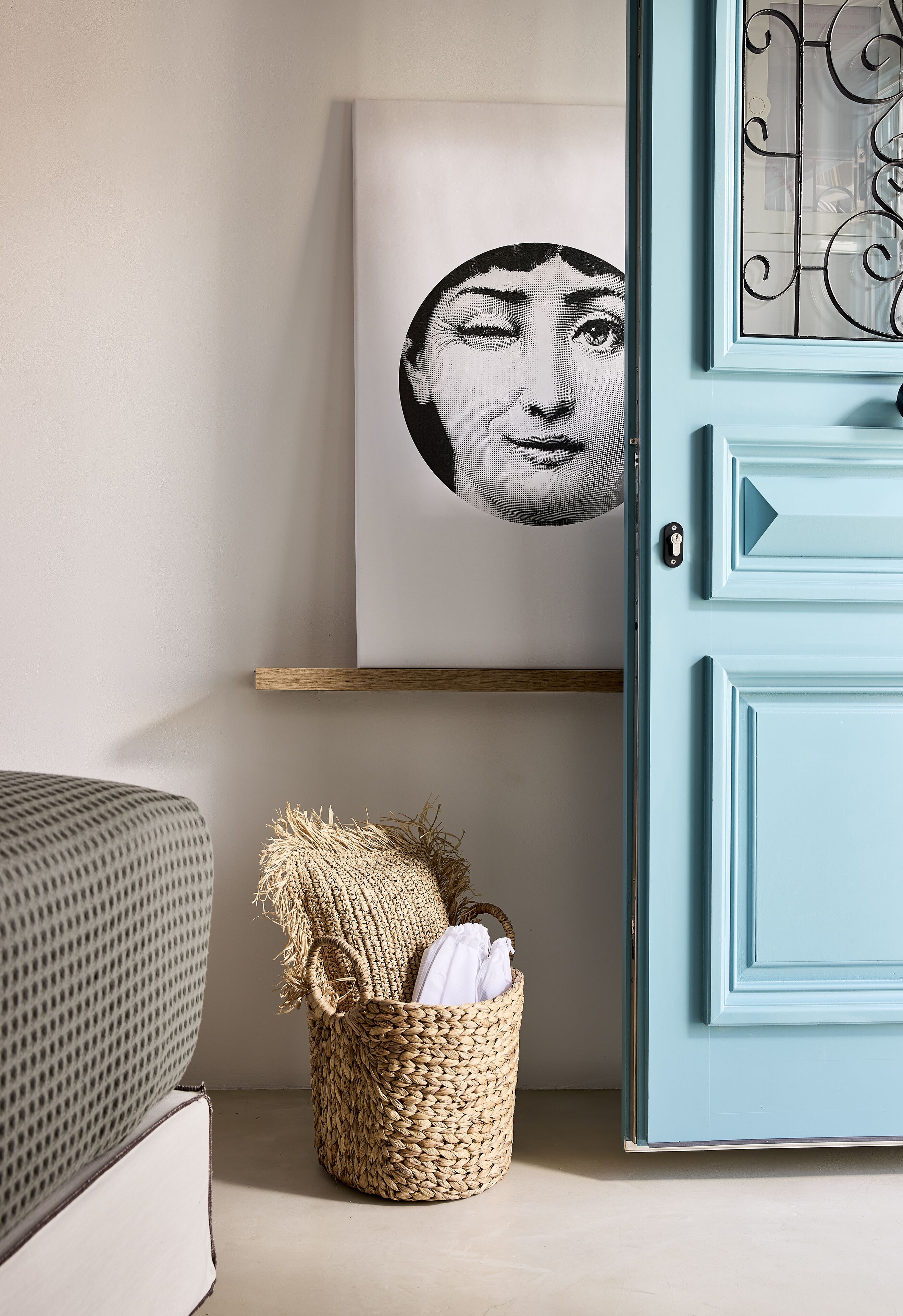 Ein Zimmer mit einer hellblauen Tür, einem gewebten Korb mit weißem Stoff und einem gerahmten Kunstwerk an der Wand.