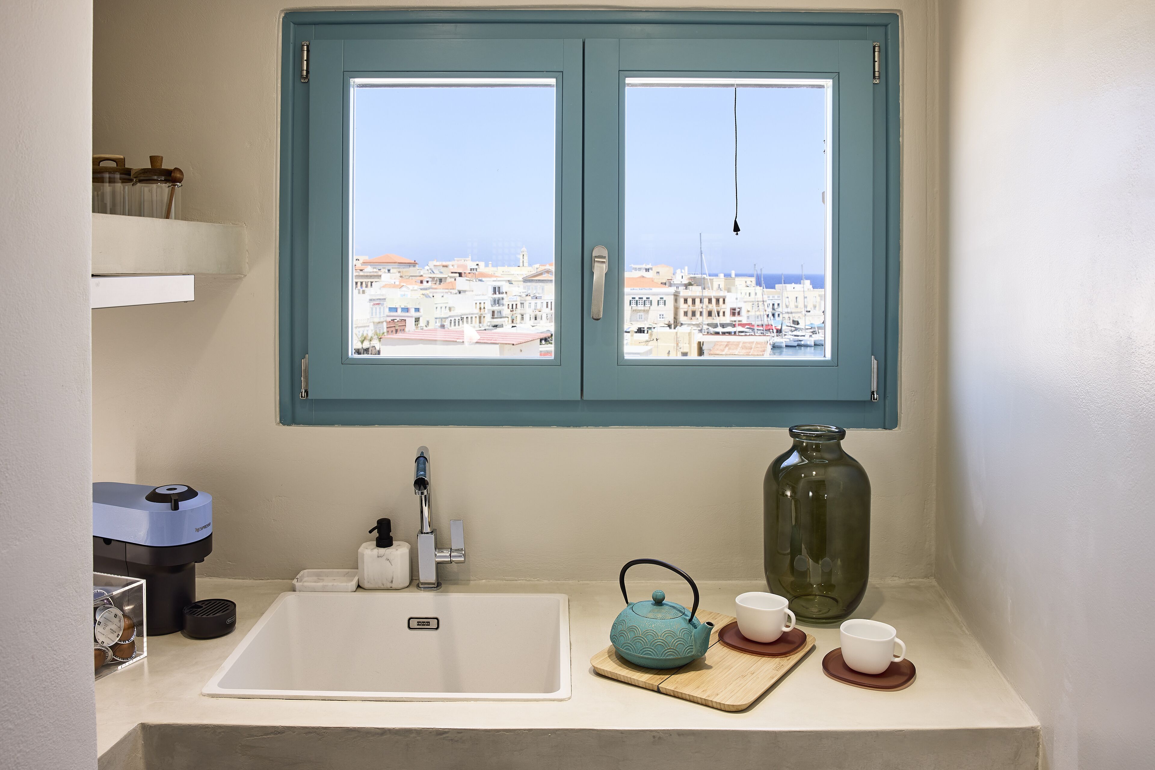 Un baño con lavamanos, una ventana con vista a edificios, un florero verde y una tetera.