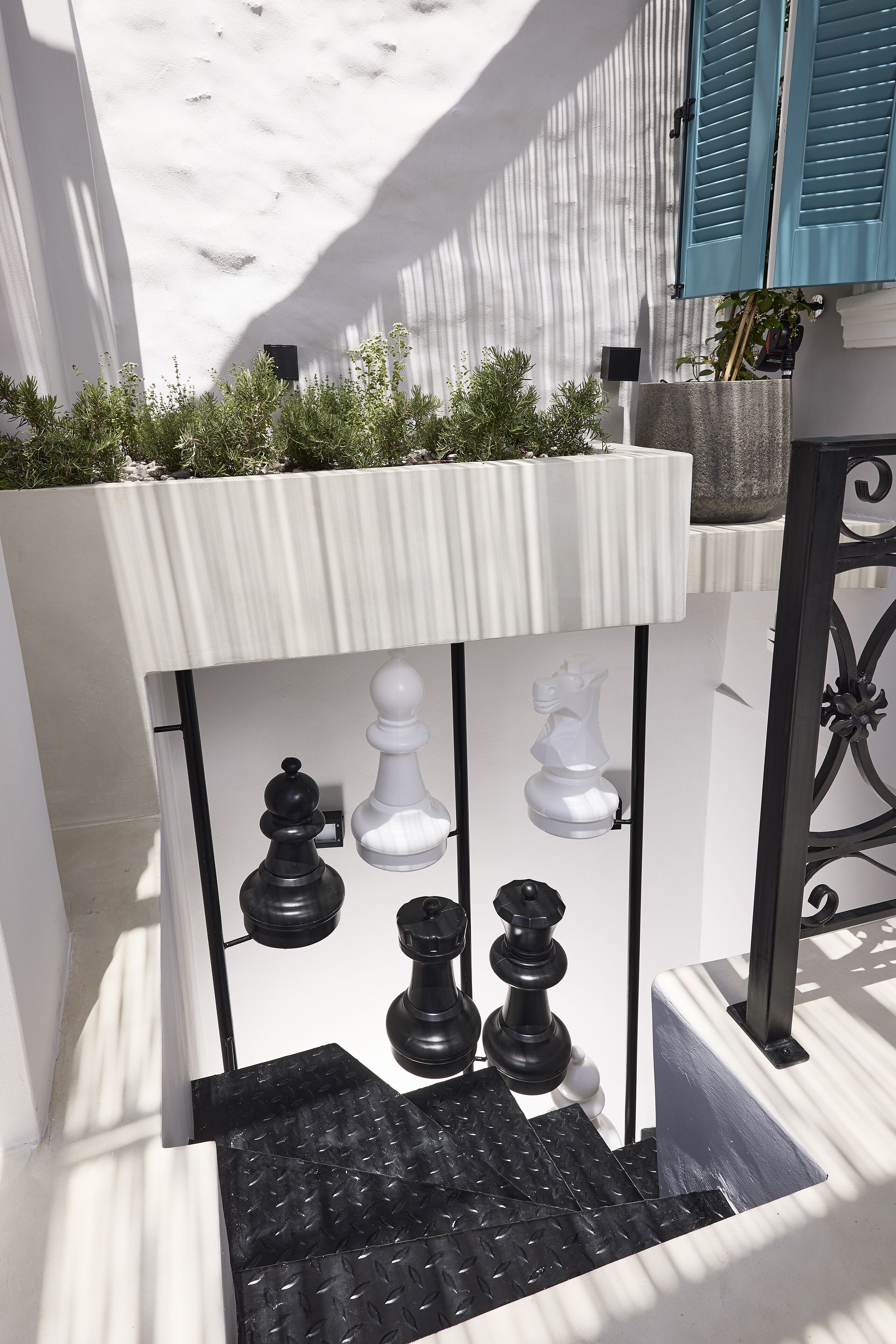 Un espace extérieur étroit, avec un mur blanc à la texture particulière, un portail en métal noir et un petit bac à fleurs contenant des plantes vertes.