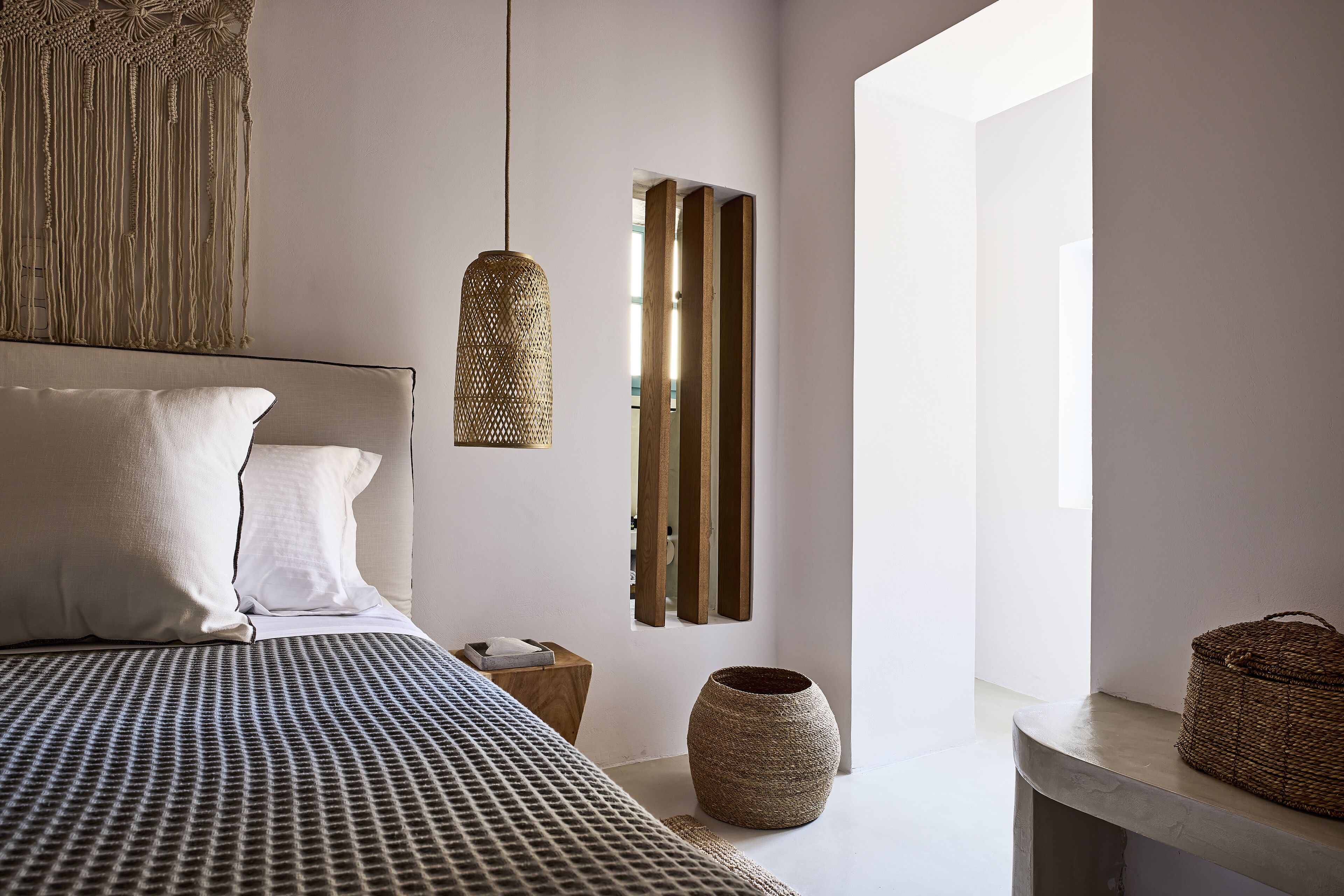 A modern bedroom with a large bed, a hanging light fixture, a wooden nightstand, a woven basket, and a large vase.