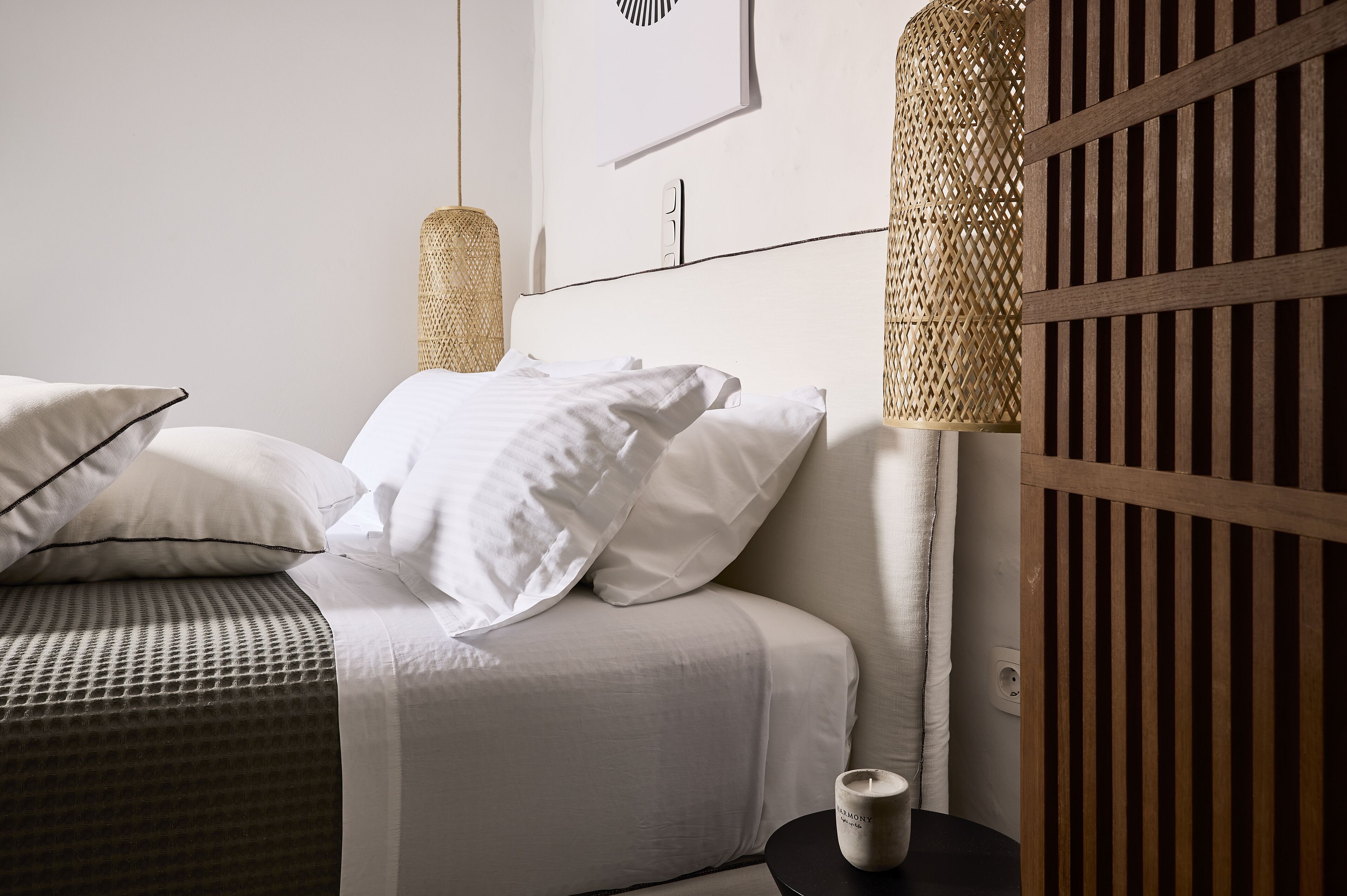Una cama bien tendida con sábanas blancas, un tapiz de pared tejido y una mesita de noche con una vela.