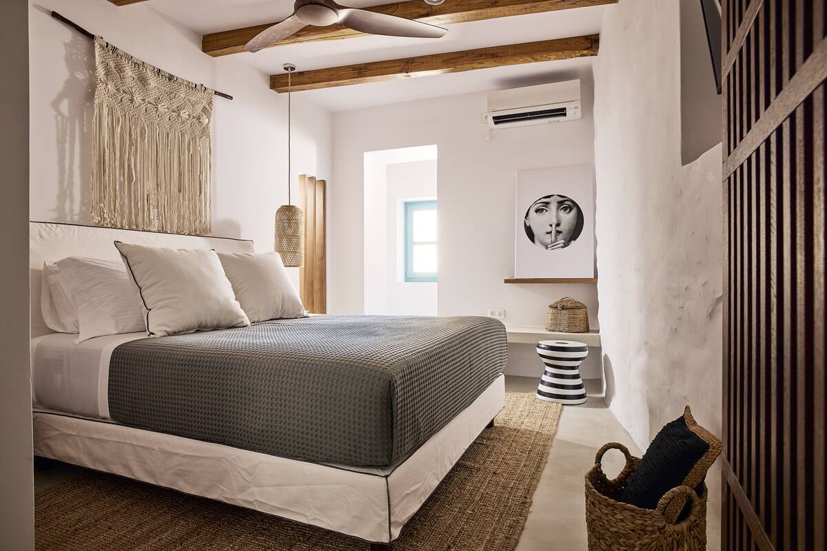 Un dormitorio con una cama, un adorno de macramé en la pared, un ventilador de techo, un aire acondicionado instalado en la pared y una ventana.
