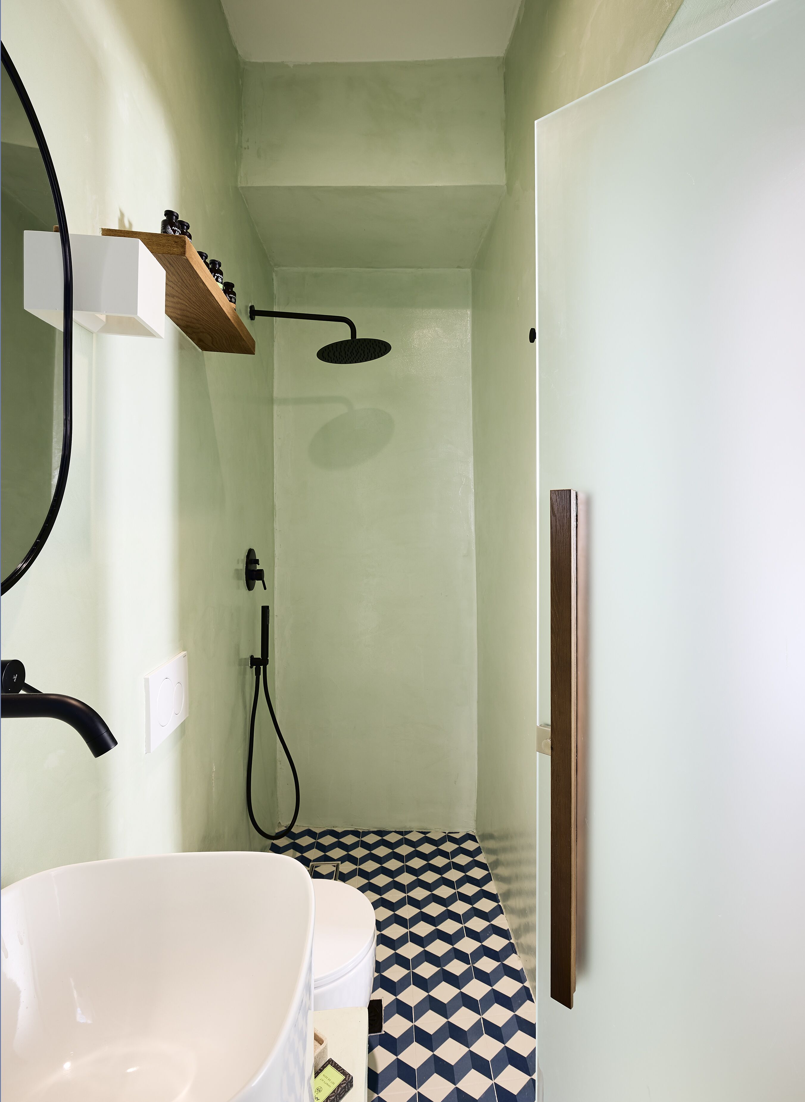 A bathroom with a white bathtub, a shower with a black hose, a wooden shelf with toiletries, and a patterned floor.