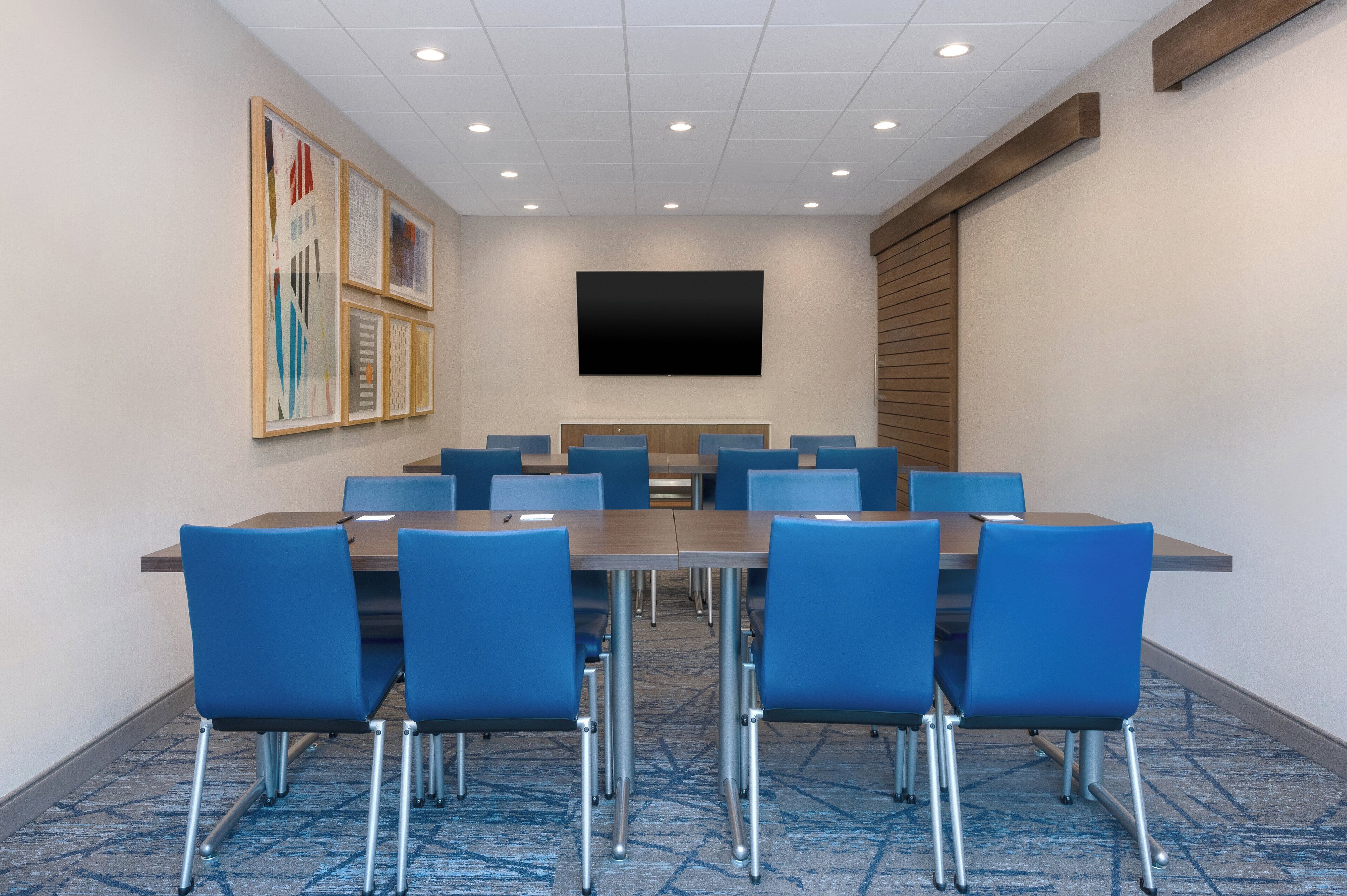 Sala de conferências com uma mesa comprida e cadeiras azuis, uma TV de tela plana na parede e quadros na parede.