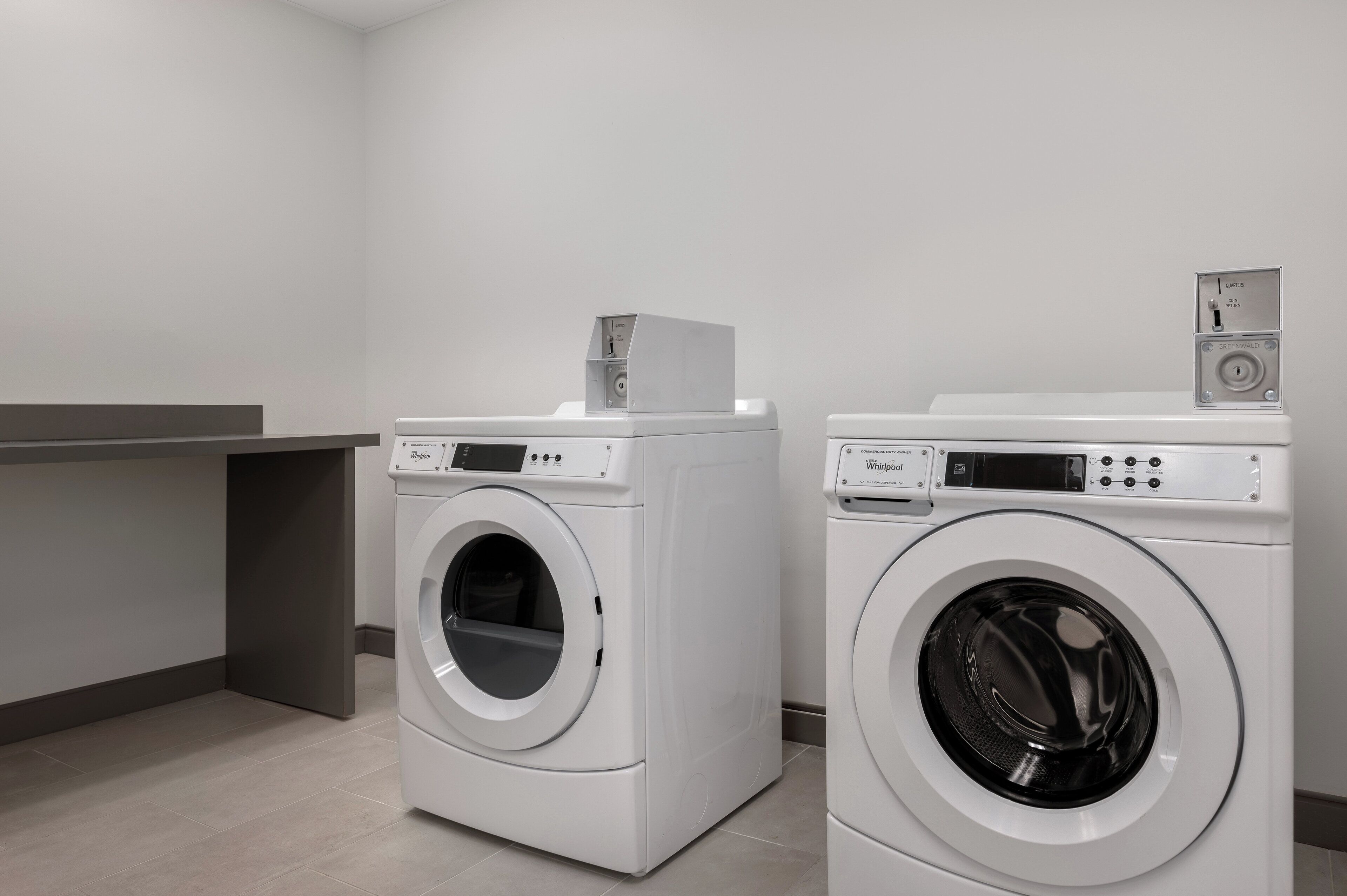 Two front-loading Whirlpool washers in a modern laundry room with a grey countertop and a grey wall.