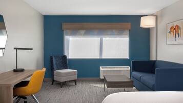 A hotel room with a wooden desk, a yellow chair, a blue sofa, a white bed, a window with blinds, a standing lamp, and a radiator.