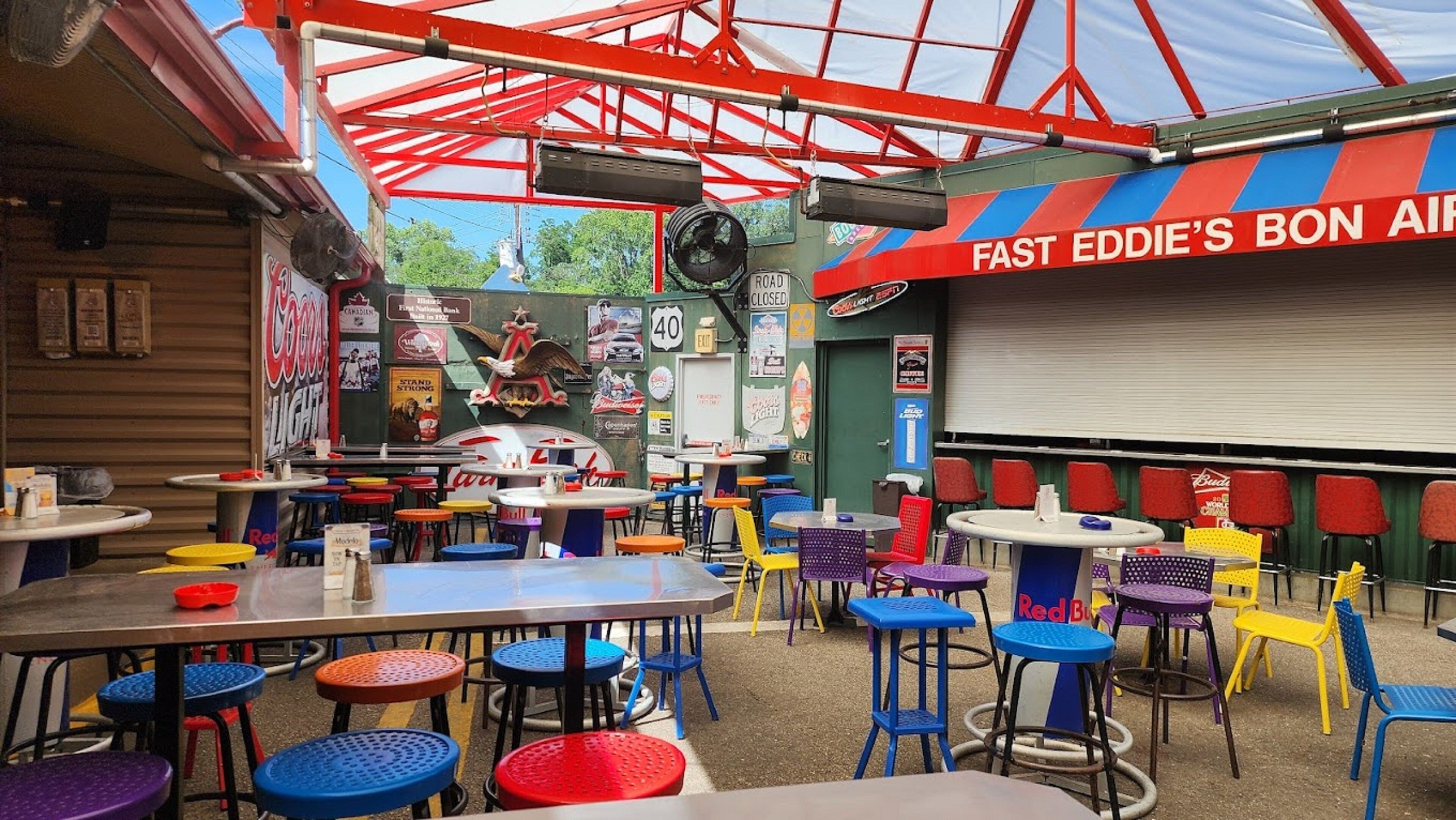 An outdoor dining area with colorful tables and chairs, a large mural, and a sign reading "FAST EDDIE'S BON AIR".