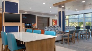 A modern indoor space with a long white table, blue and orange chairs, a sofa with patterned cushions, and a coffee station. - Holiday Inn Express and Suites Alton St Louis Area by IHG (Alton)