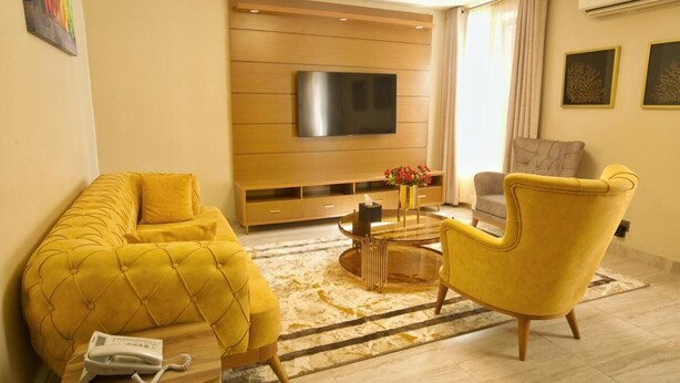 A modern living room with a yellow sofa, a yellow armchair, a glass coffee table, and a flat-screen TV mounted on a wooden wall. - MUSADA LUXURY HOTELS AND SUITES (Abuja)