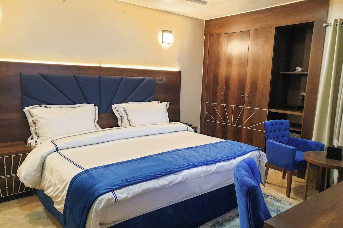 A modern hotel room with a large bed, a wooden wardrobe, a blue chair, and a small table.