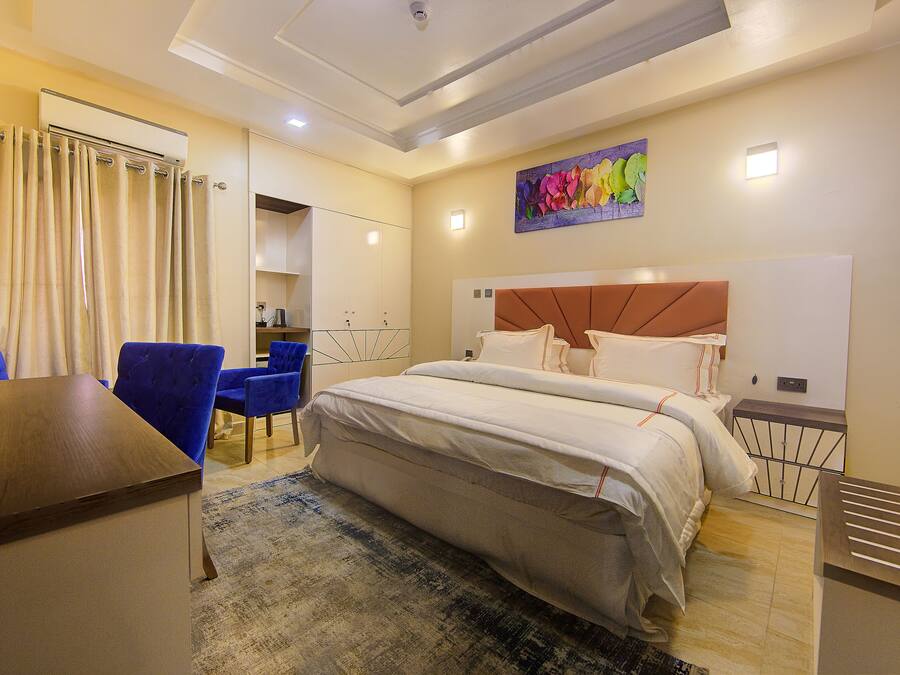A hotel room with a bed, a desk, and a chair. There is a painting on the wall and a window with curtains.