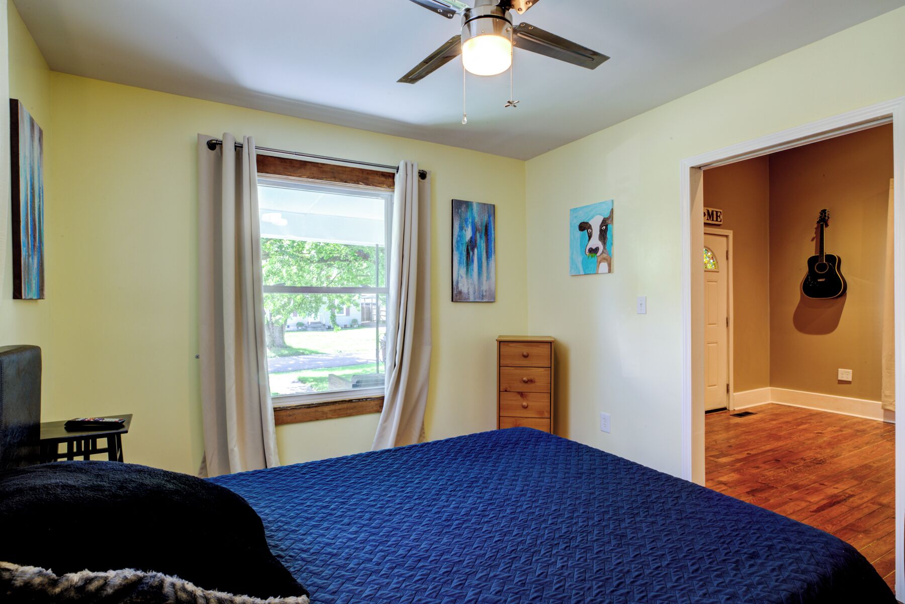 Une chambre à coucher avec un lit, un ventilateur de plafond, une fenêtre avec des rideaux, une commode en bois et une guitare accrochée au mur.