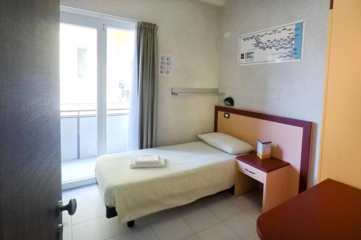 Une chambre d’hôtel avec un lit simple, une table de chevet, une petite table, une fenêtre avec des rideaux et une carte accrochée au mur.