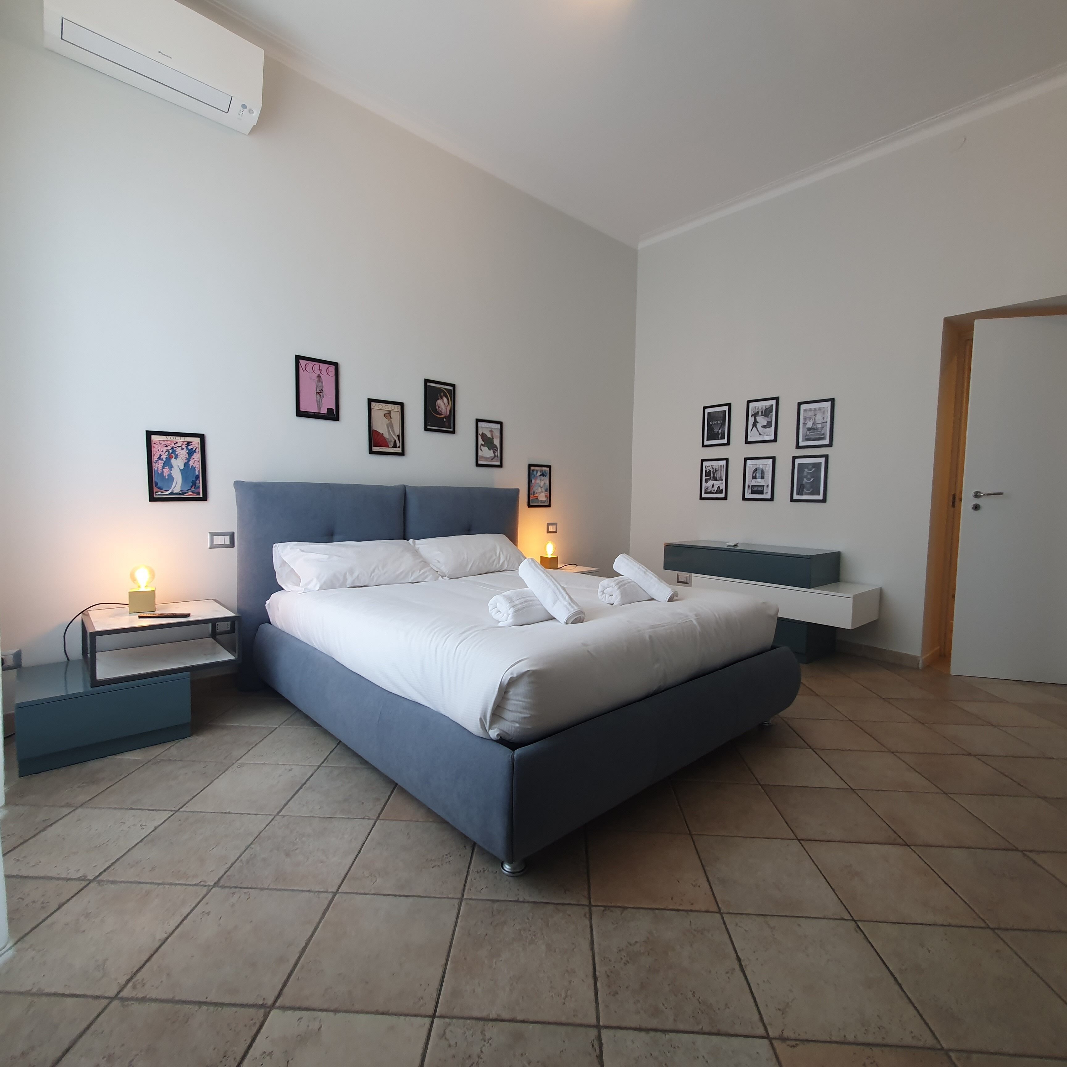 A modern bedroom with a large bed, bedside tables, a bedside lamp, and a wall-mounted air conditioner.