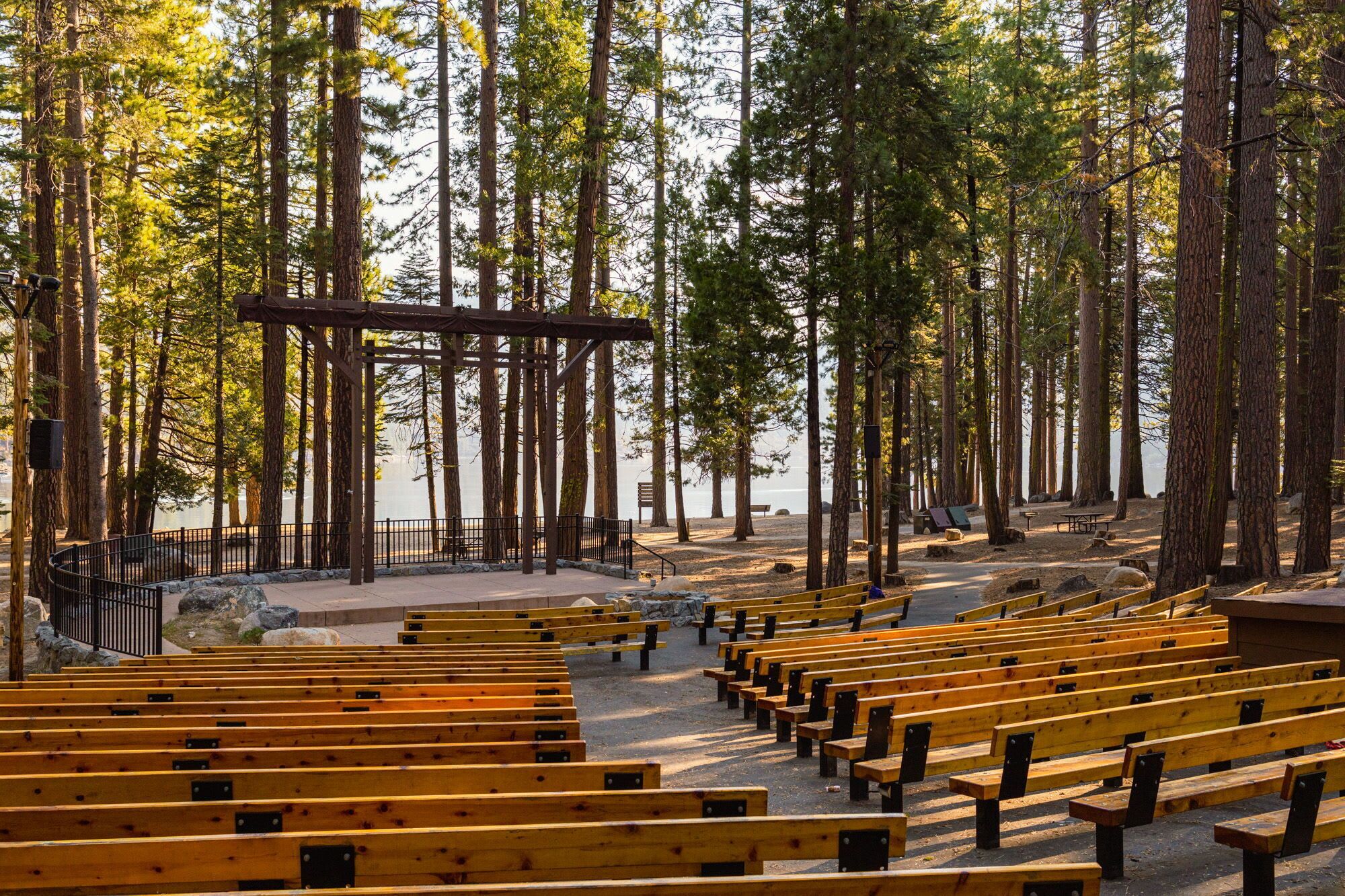 Une zone boisée avec des bancs en bois disposés en rangées, une pergola en métal et une vue dégagée sur les arbres environnants.