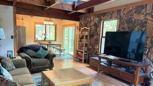 A cozy living room with a stone wall, wooden furniture, a television, and a dining area with natural light. - Pinecrest Escape (Cold Springs)