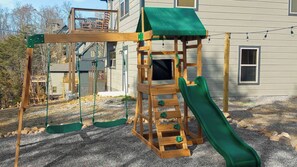 A wooden play structure with a slide and climbing elements in front of a house.