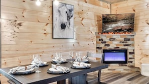 A dining area with a long table set for six, a fireplace with a stone surround, and a mounted television. - Cabin Bliss | Hot Tub, Games & Scenic Yard Escape (Sevierville)